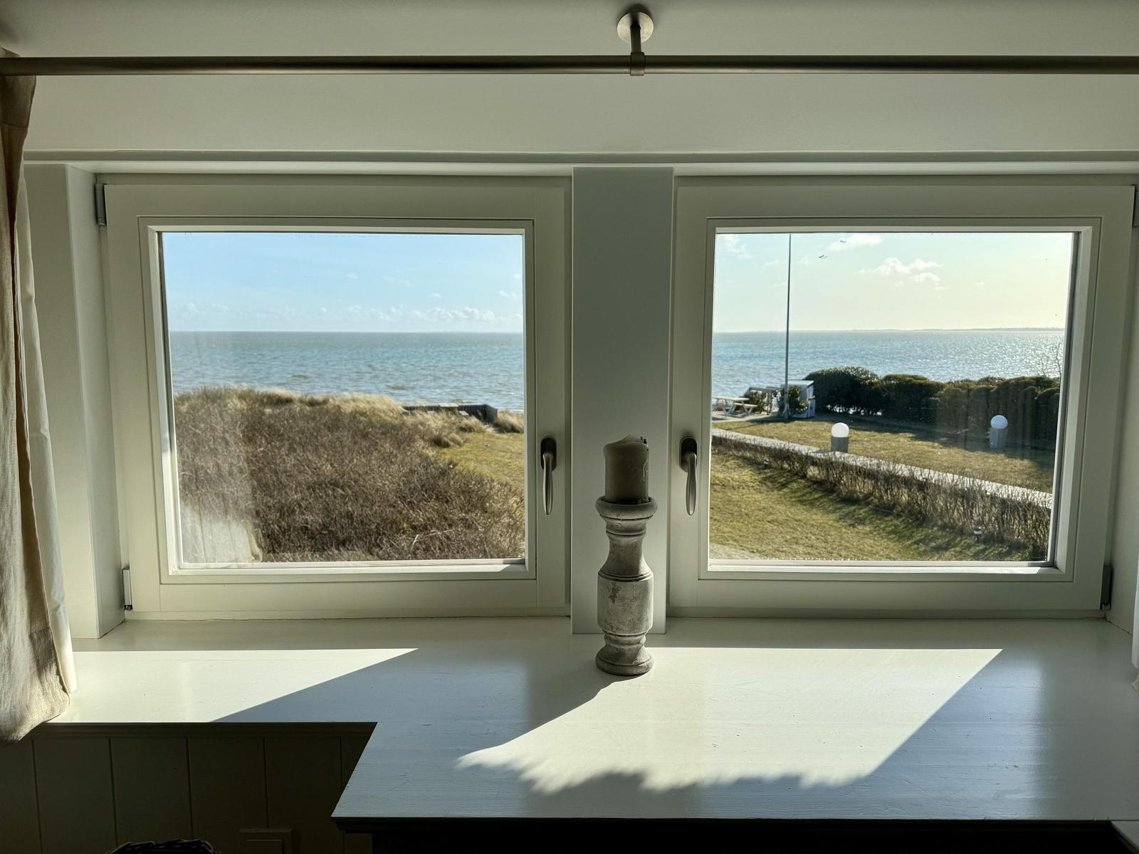 Zwei Fenster mit Blick auf das Meer und Grünflächen. Auf dem Fensterbrett steht eine Kerze.