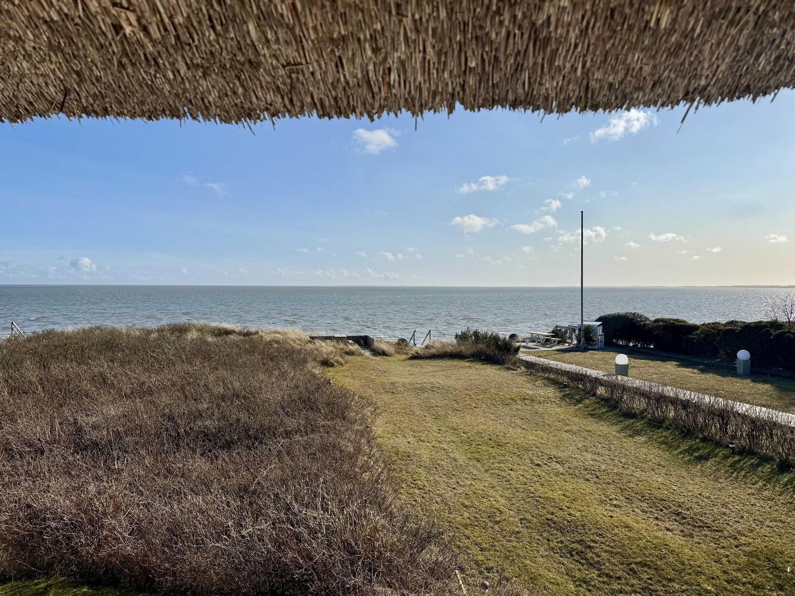 Weitläufiger Blick auf das Meer unter einem strohgedeckten Dach mit Rasenfläche und Sträuchern.