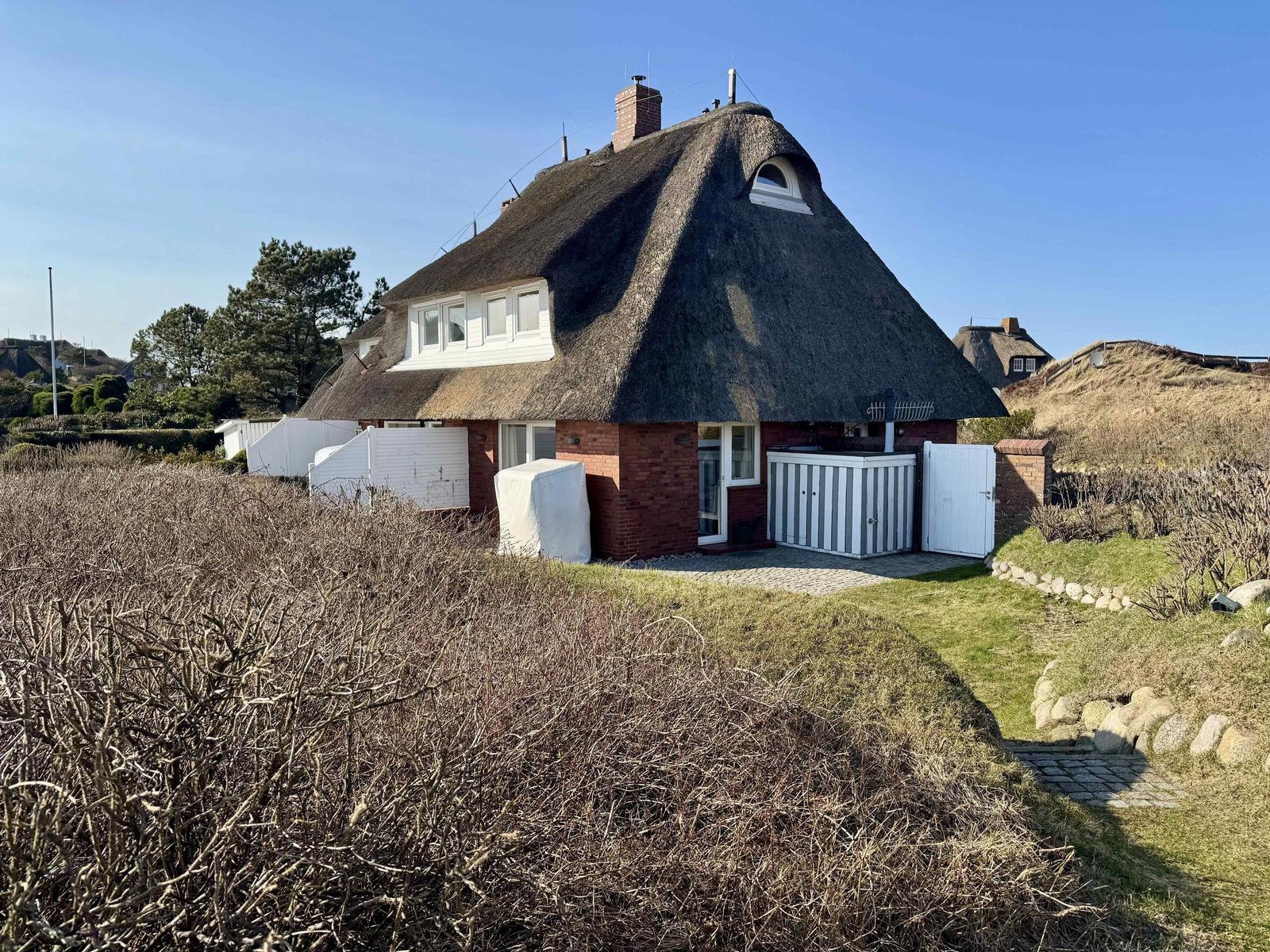 Dorfhaus mit Strohdach, weißen Fenstern und rotem Mauerwerk. Garten mit Hecken und Steinen.