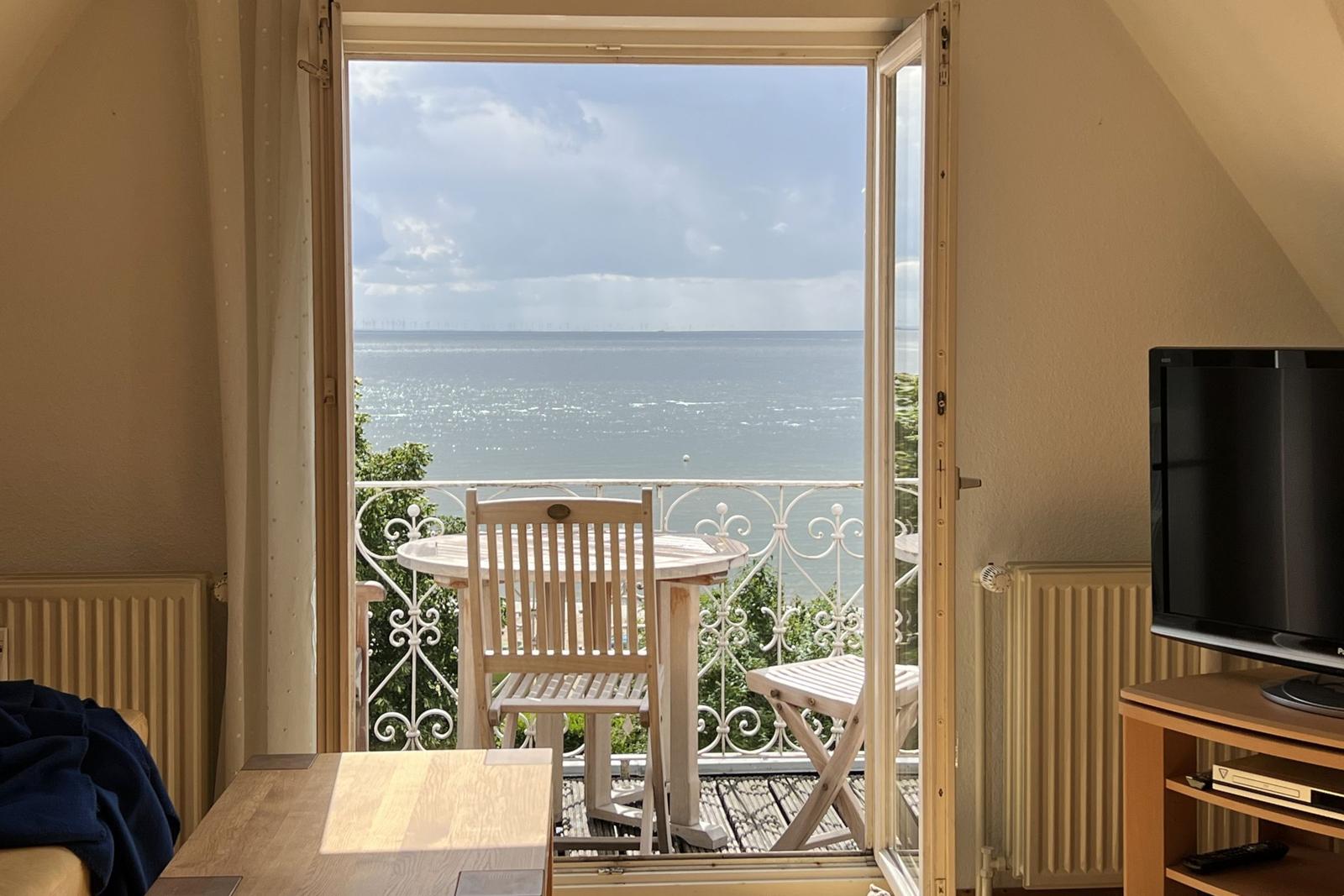Durch die offene Tür sieht man einen Balkon mit Tisch und Stühlen. Dahinter liegt das Meer.