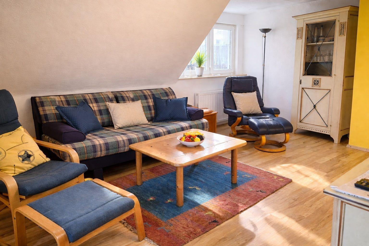 Wohnzimmer mit Sofa, Sessel, Tisch und Teppich. Fenster, Schrank und Holzboden.