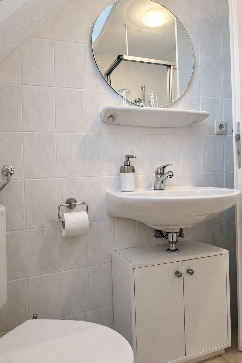 Badezimmer mit Waschbecken, Spiegel und Toilette. Wandfliesen und weißer Schrank.