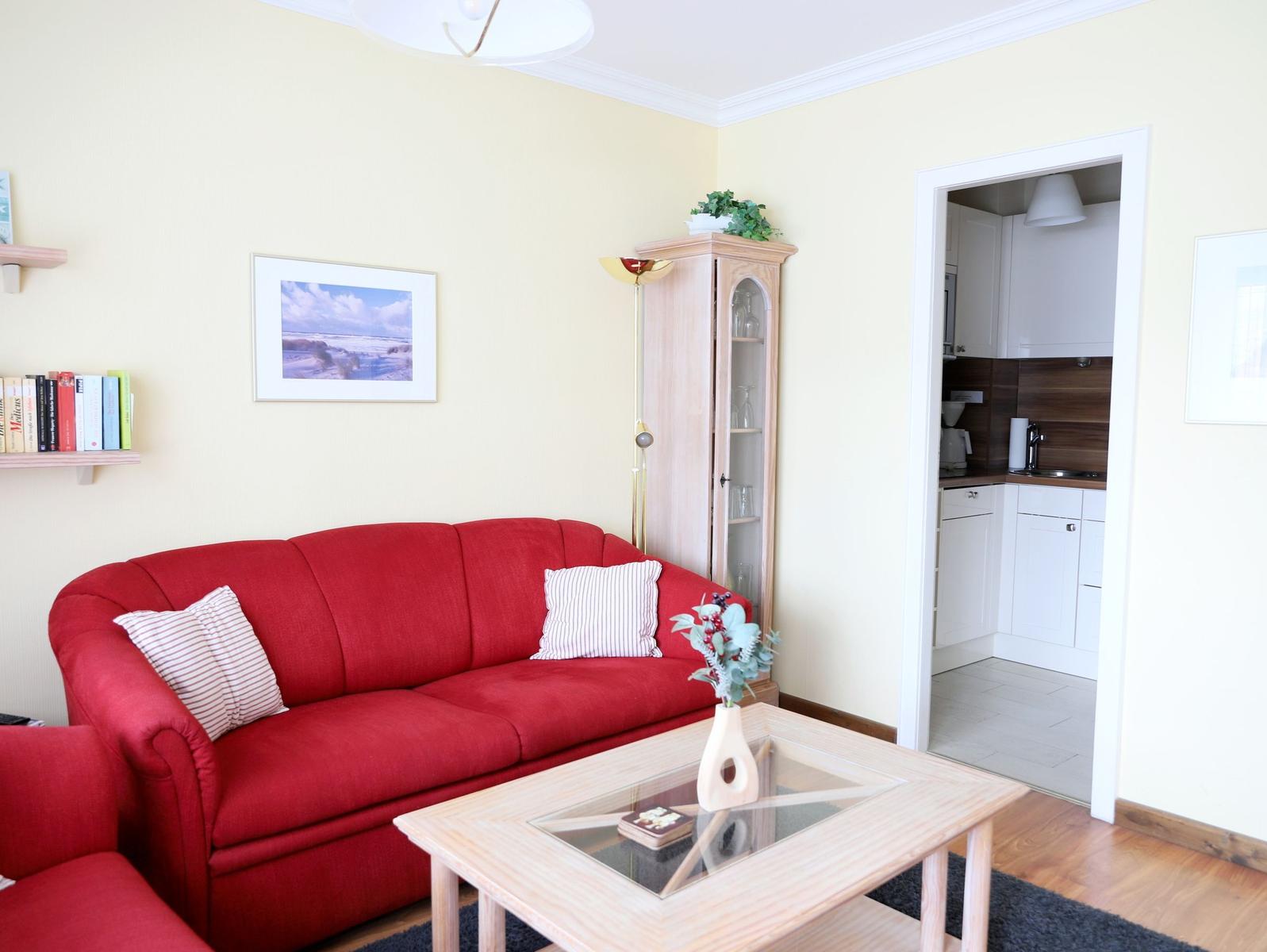 Ein Wohnzimmer mit rotem Sofa, Glas-Tisch und Blick in die Küche.