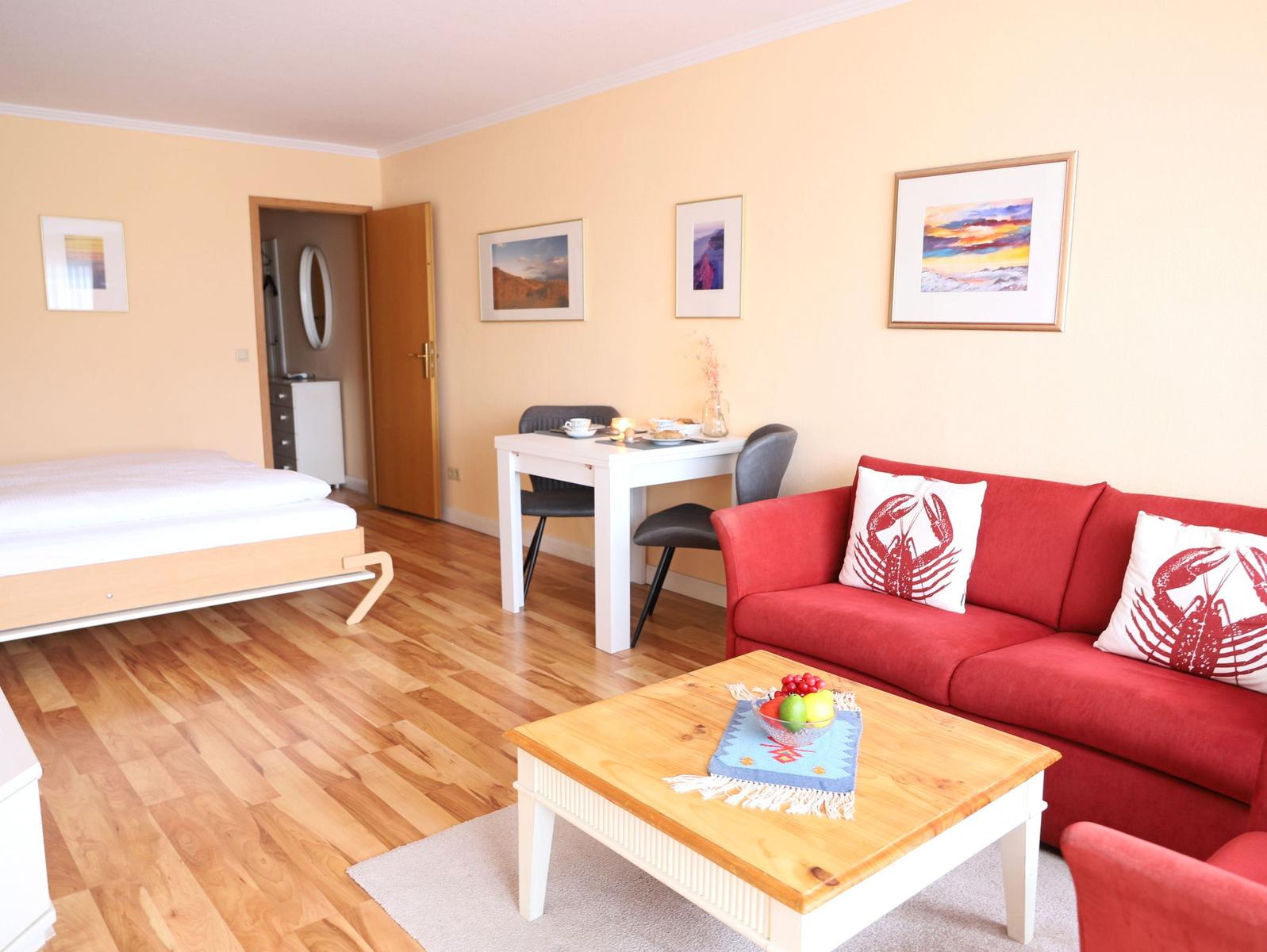 Ein Wohnzimmer mit rotem Sofa, Esstisch und Bett. Holzboden und Wandbilder.