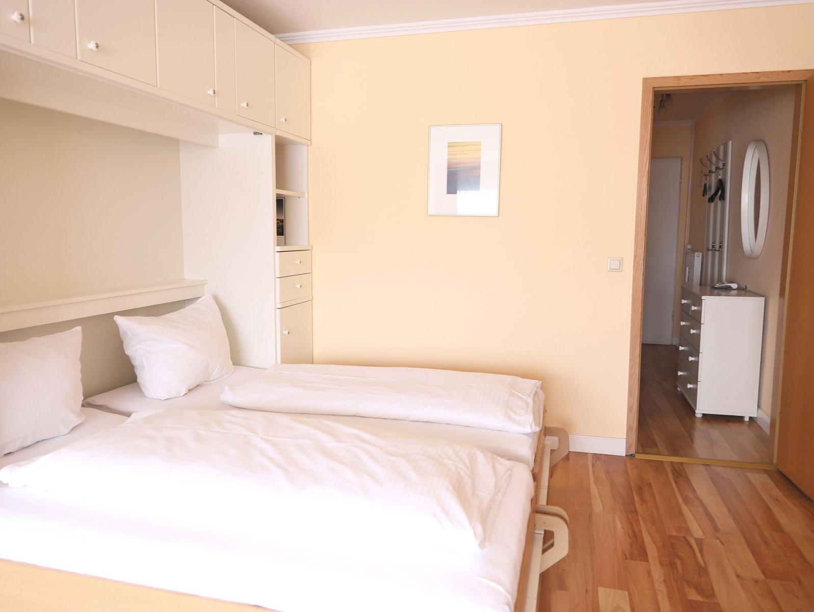 Schlafzimmer mit Doppelbett, weißen Laken und integriertem Schrank. Holzfußboden und angrenzender Flur.