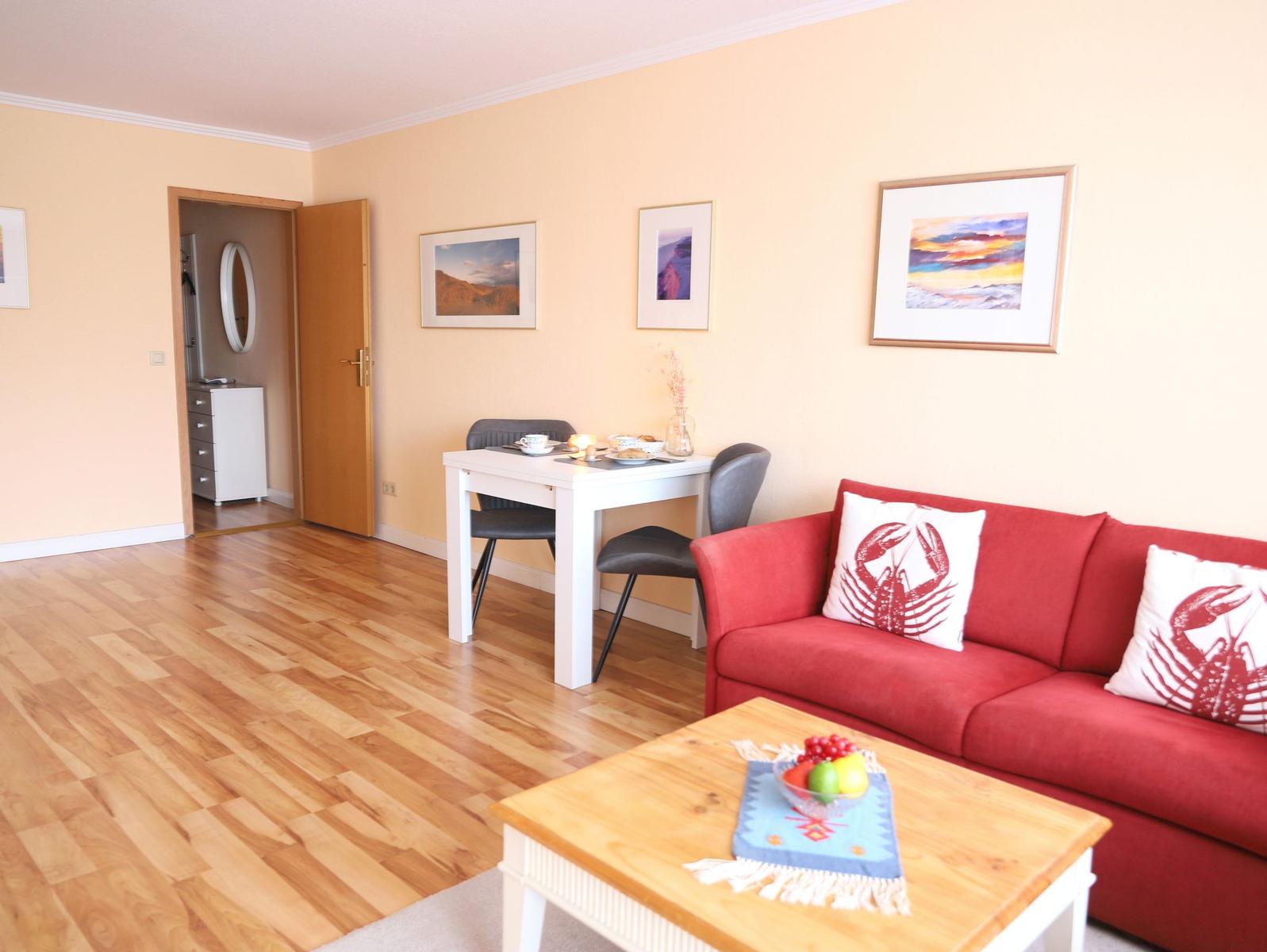 Wohnzimmer mit rotem Sofa, Esstisch und Holzboden. An der Wand hängen Bilder.