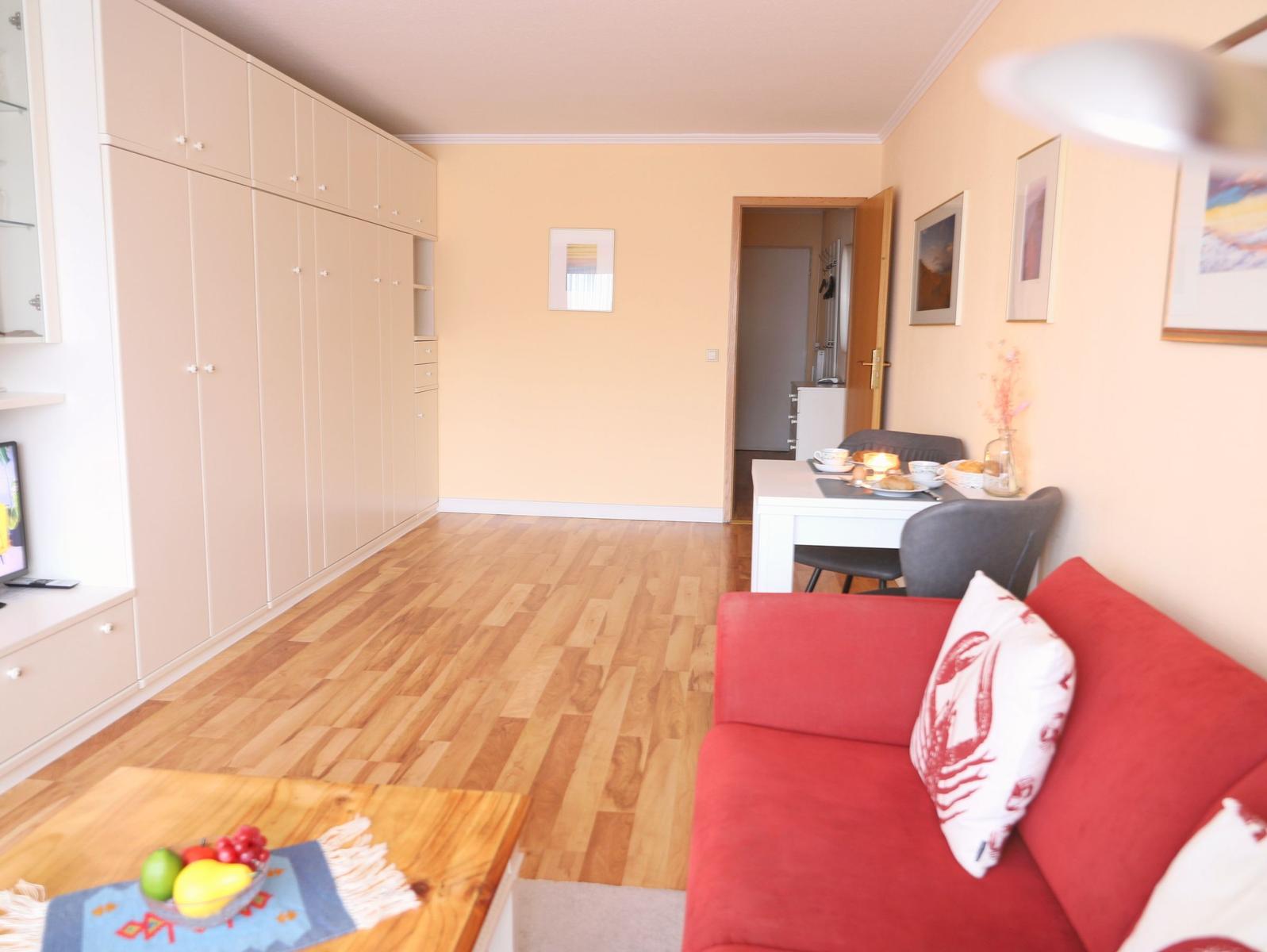 Wohnzimmer mit rotem Sofa, Holzboden und integriertem Schrank. Essbereich mit Tisch und Stühlen.
