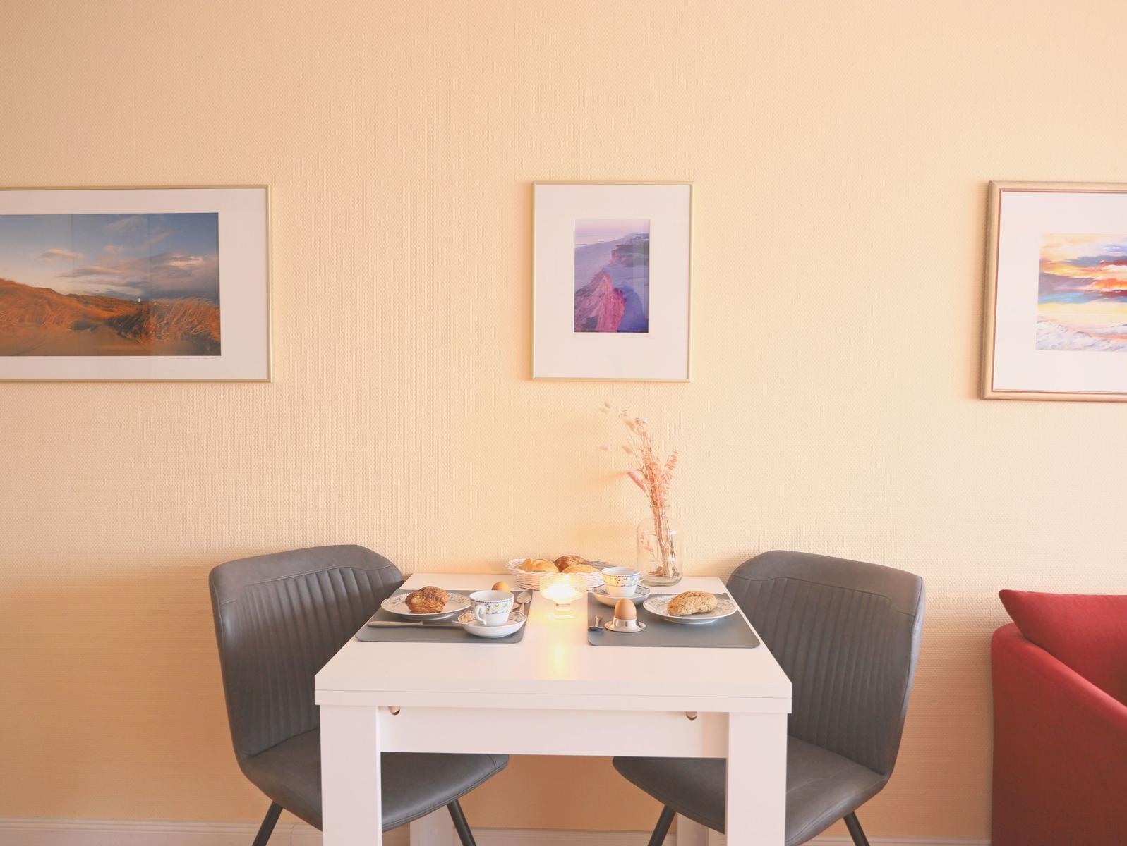 Esstisch mit zwei Stühlen, Wandbilder und einer roten Couch im Hintergrund.