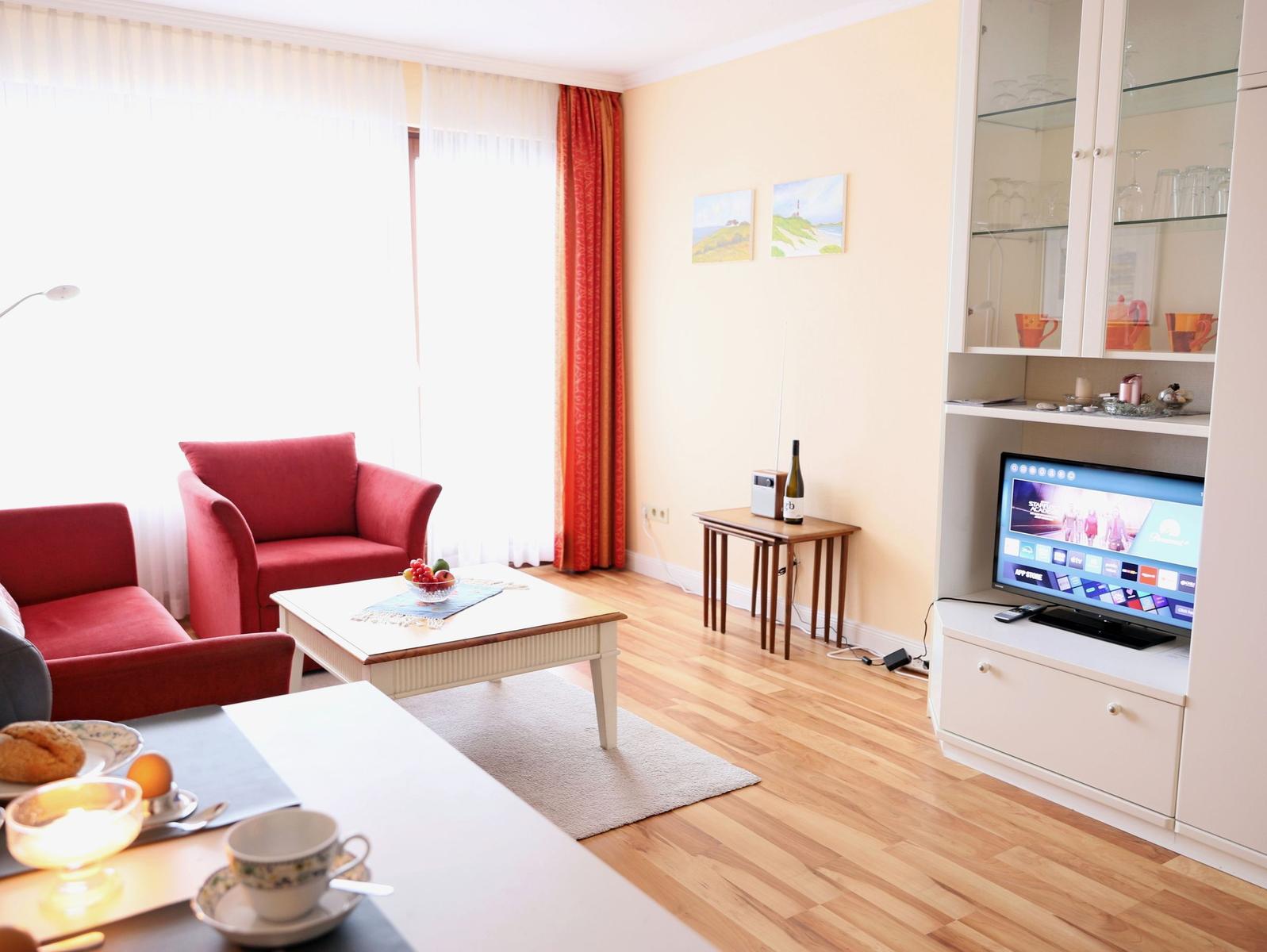 Wohnzimmer mit rotem Sessel, TV und Holzboden. Fenster mit Vorhängen.