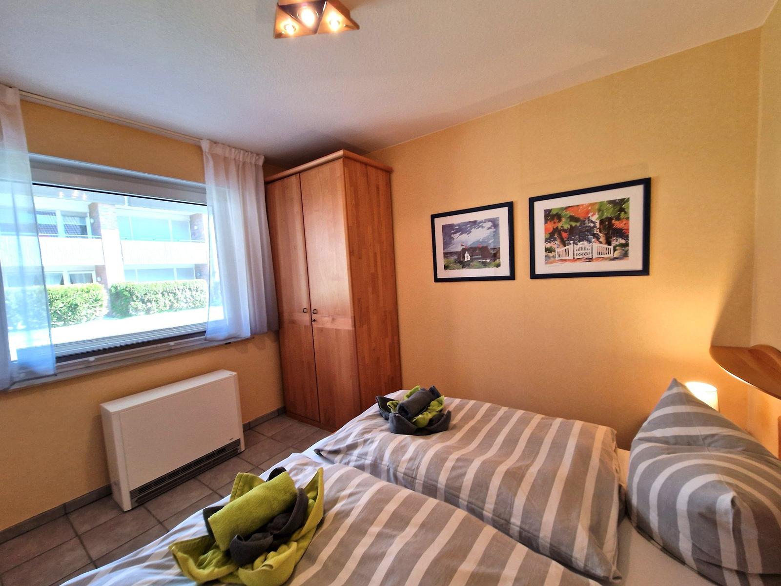 Schlafzimmer mit Bett, Kleiderschrank und Fenster. An der Wand hängen zwei Bilder.
