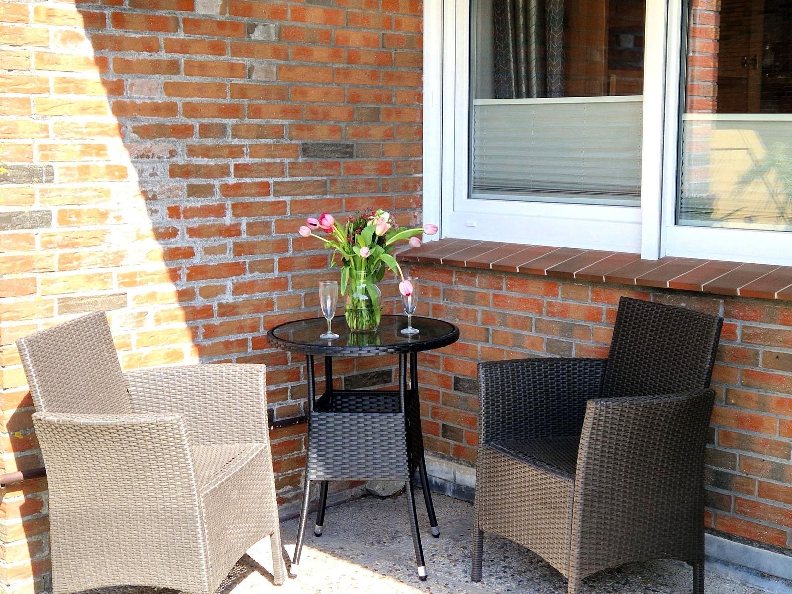 Terrasse mit Tisch und zwei Stühlen, Blumen und Gläser auf dem Tisch.