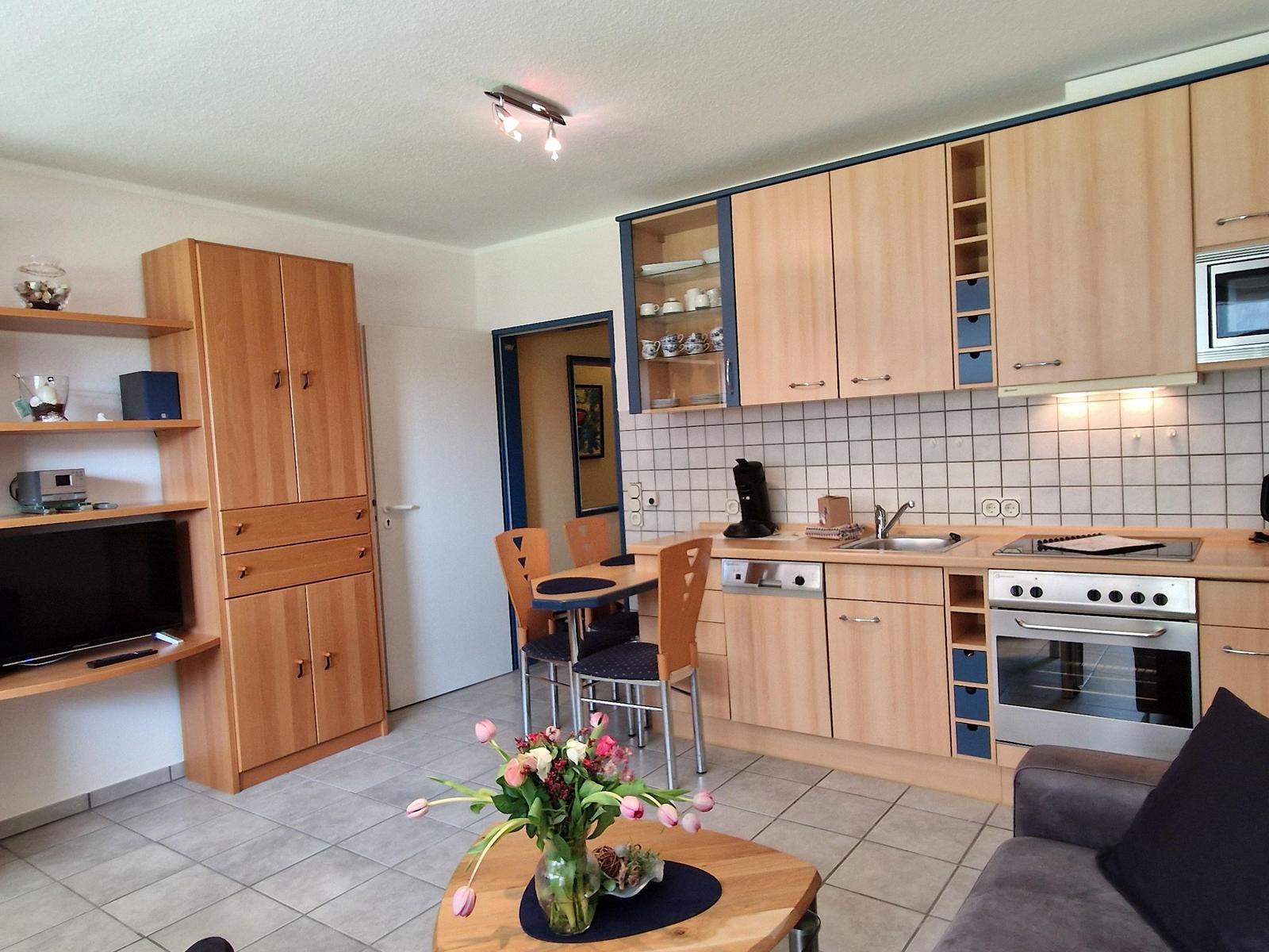 Küche mit Holzschränken, Herd und Spüle. Essbereich mit Tisch und Stühlen. Wohnbereich mit Sofa und Fernseher.