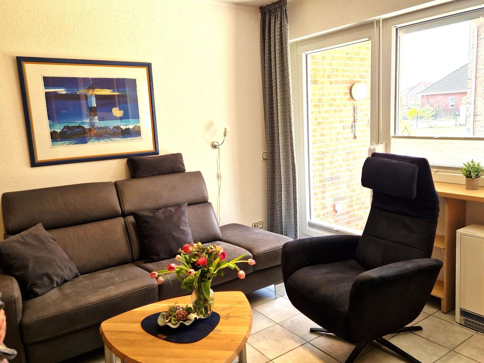 Ein Wohnzimmer mit Sofa, Sessel, Tisch und Blumen. An der Wand hängt ein Bild.