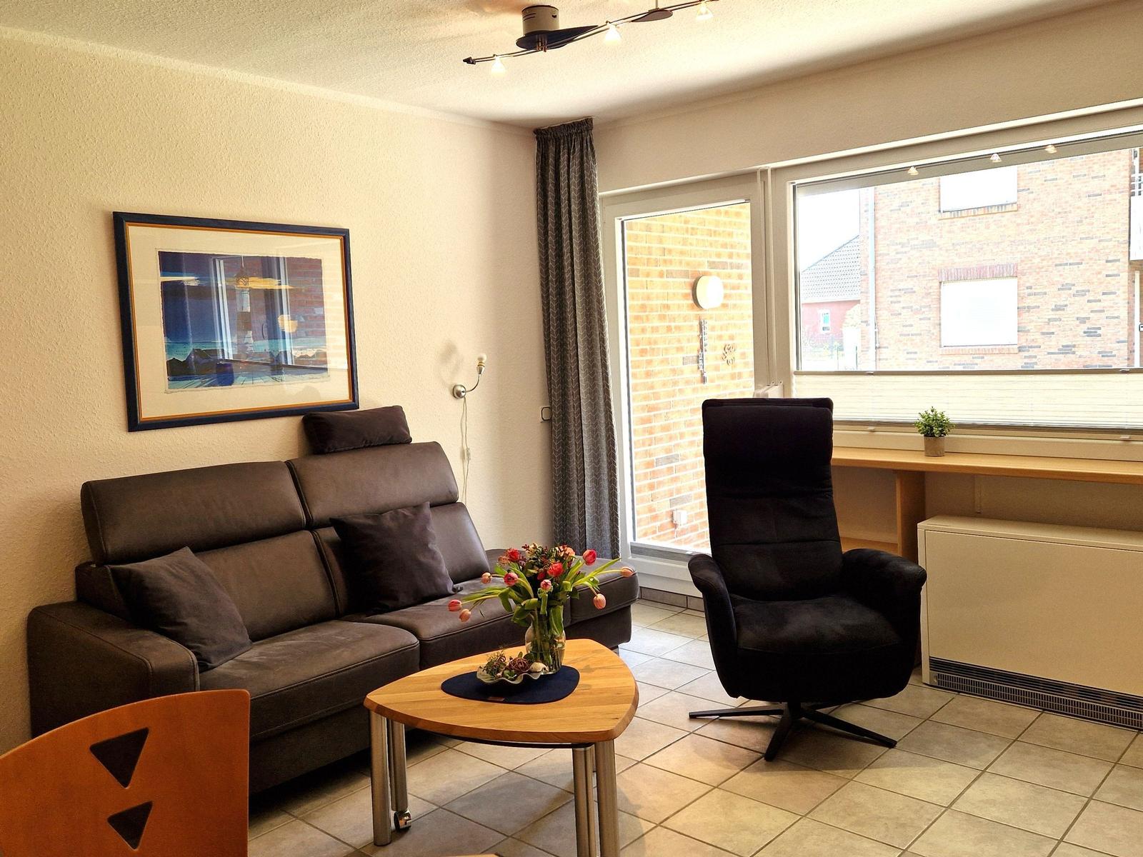 Wohnzimmer mit Sofa, Sessel, Tisch und Fenster. Wandbild und Blumen sind sichtbar.