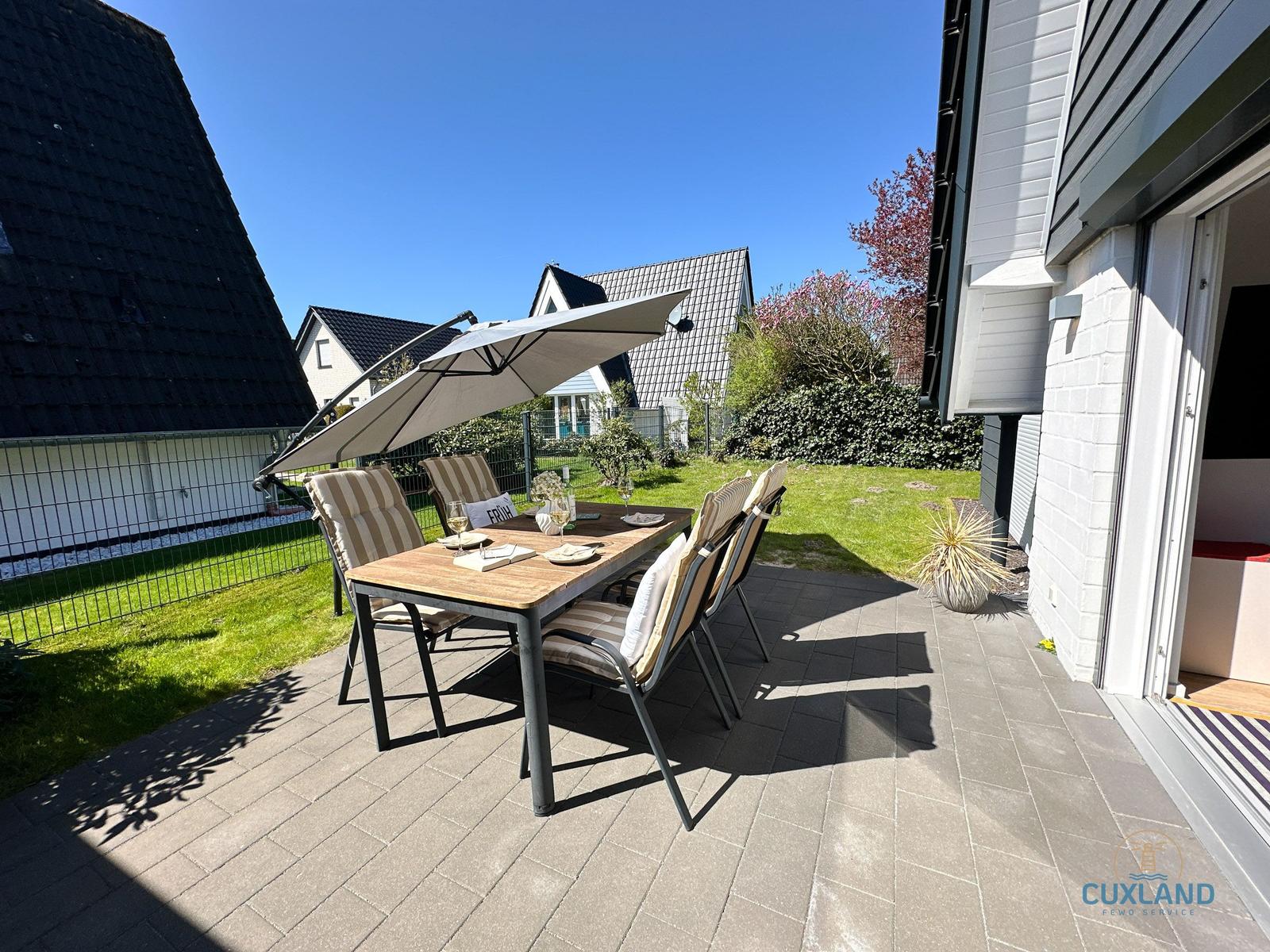 Terrasse mit Tisch, Stühlen und Sonnenschirm. Grünflächen und Häuser im Hintergrund.