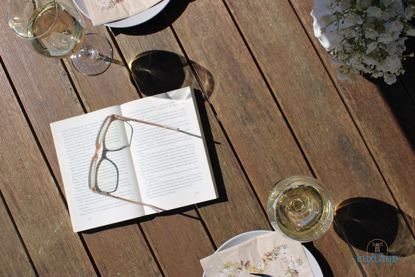 Ein Tisch mit einem Buch, einer Brille, zwei Weingläsern und Blumen.