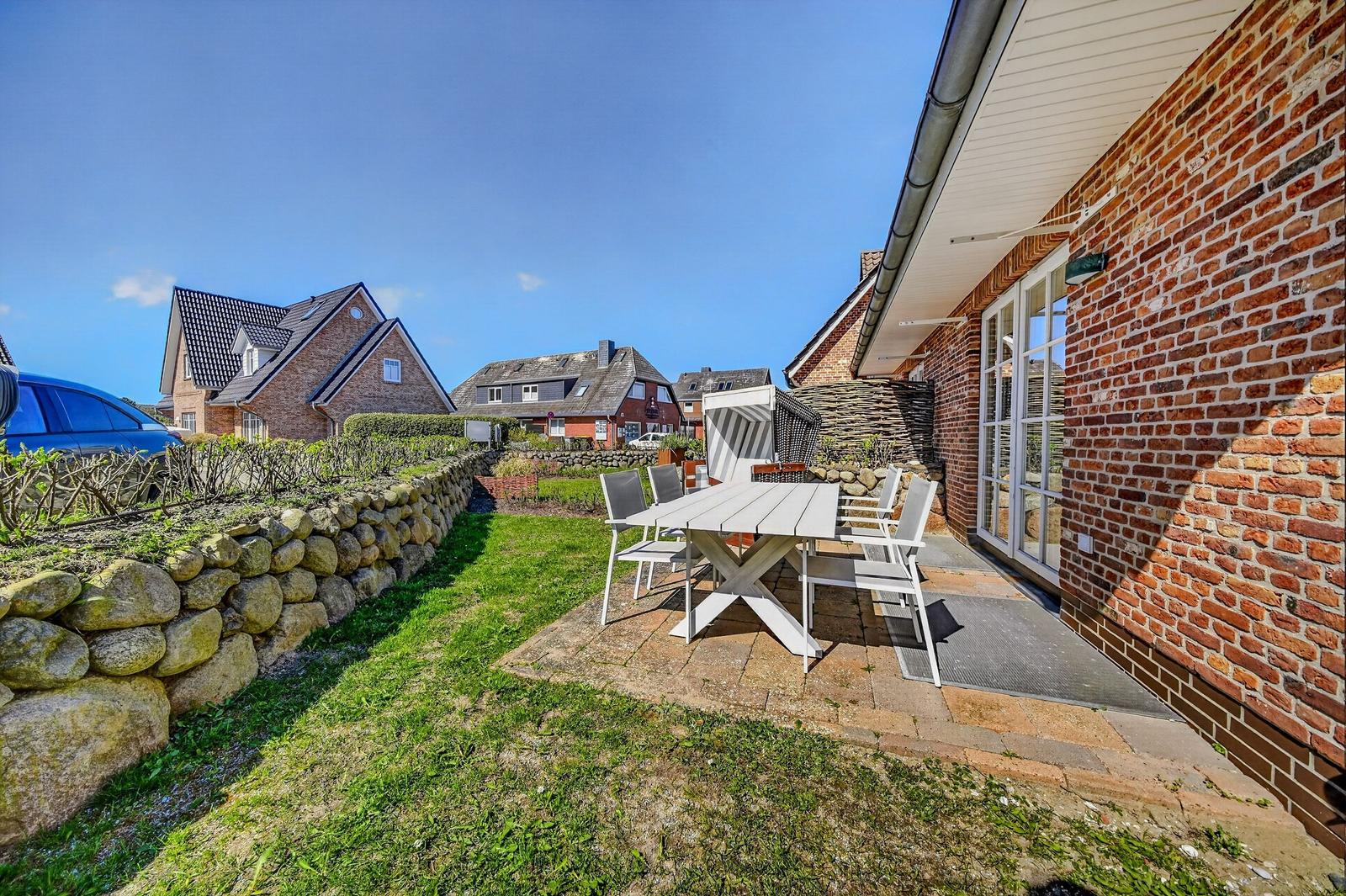 Terrasse mit Tisch und Stühlen neben Backsteinhaus. Grünfläche und Steinmauer im Hintergrund.