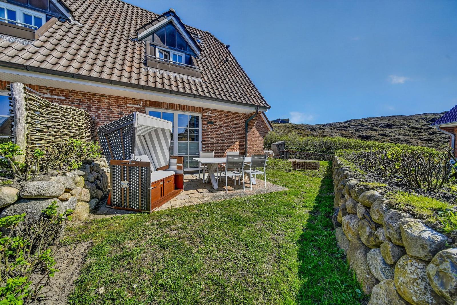 Haus mit Terrasse, Gartenmöbel und Strandkorb. Steinmauer und Grünflächen umgeben den Außenbereich.