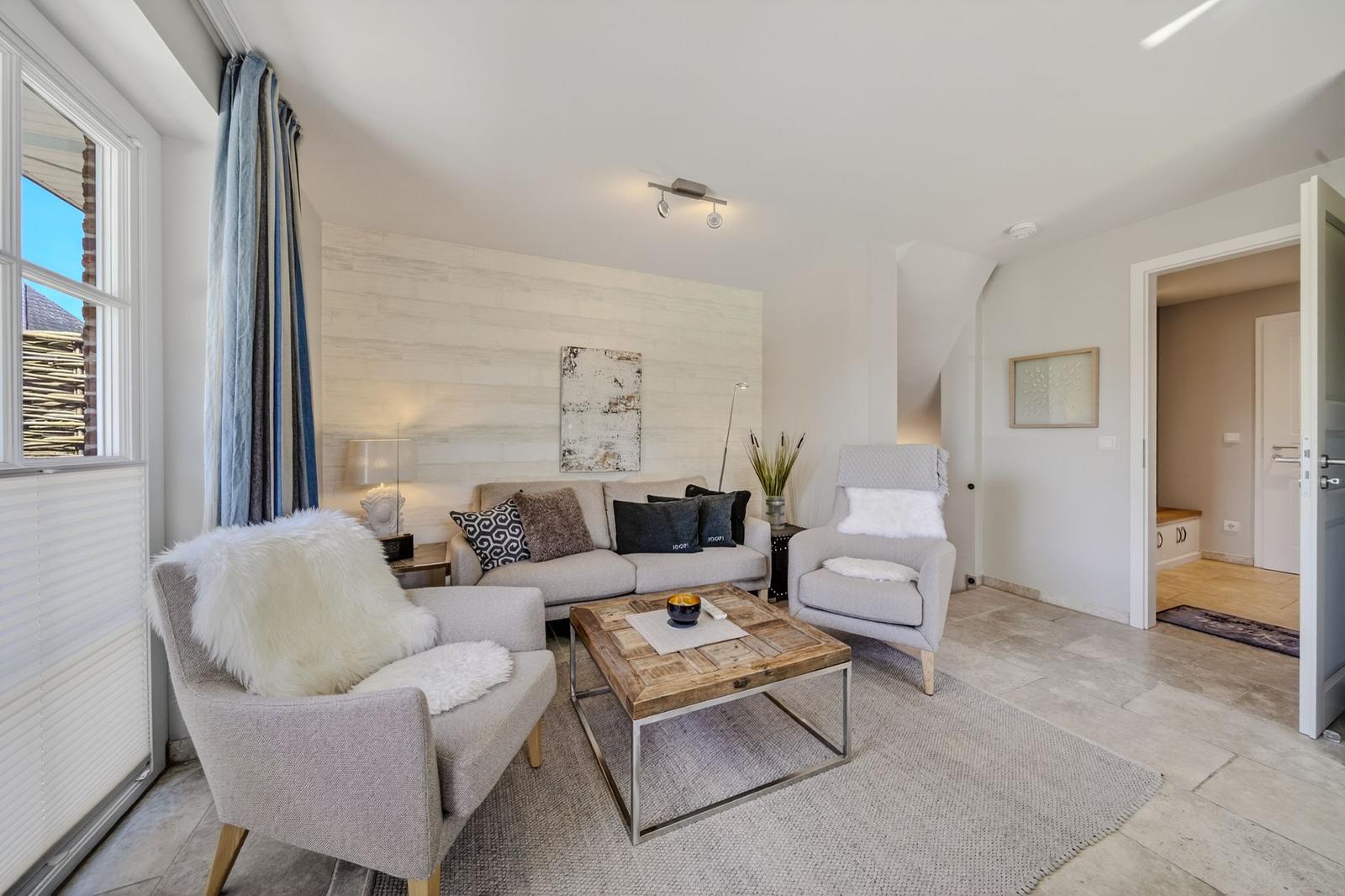 Wohnzimmer mit Sofa, zwei Sesseln, Holztablett und Fenster mit Vorhängen.