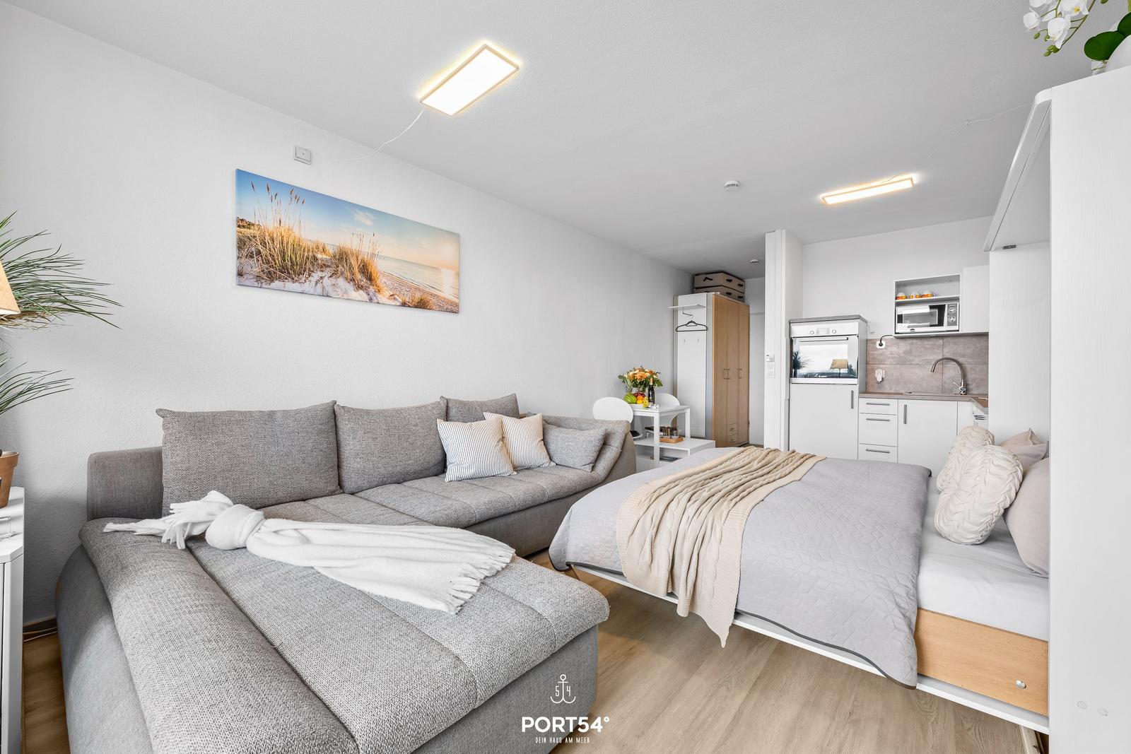 Wohnbereich mit Sofa, Bett und Küchenzeile. Wandbild zeigt Strand.