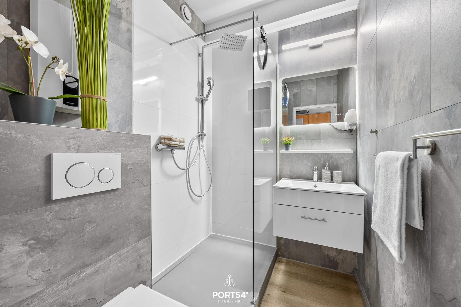 Moderne Dusche mit Glaswand, Waschbecken und Toilette in einem Badezimmer mit grauen Fliesen.