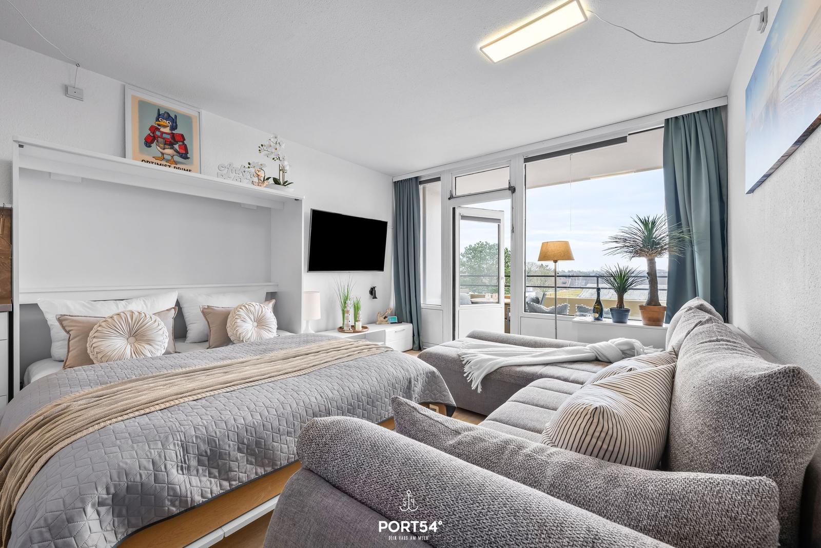 Schlafzimmer mit Bett, Sofa und TV. Balkon mit Pflanzen und Sicht auf Außenbereich.
