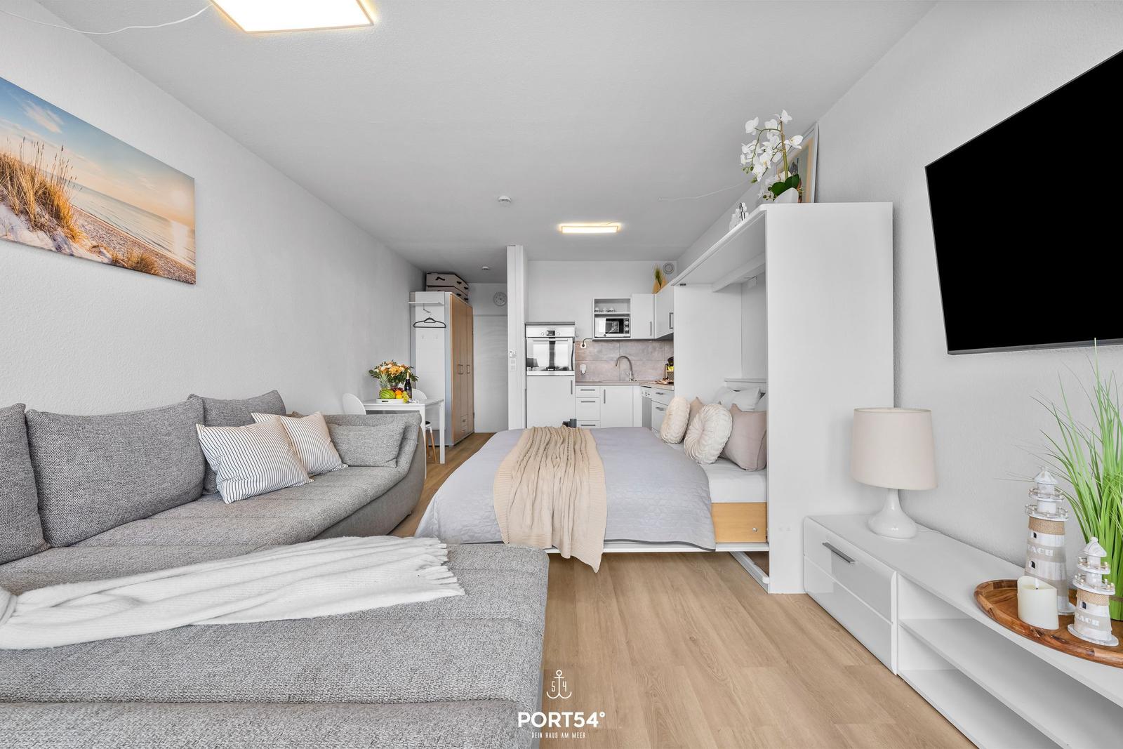 Wohnbereich mit Sofa, Bett und Küchenzeile. Wandbild und Fernseher sichtbar.