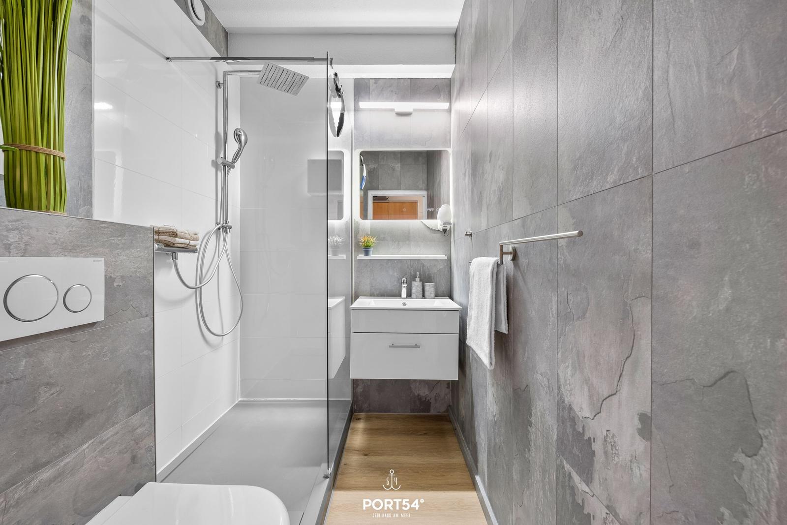 Moderne Dusche mit Glaswand, Waschbecken und Toilette in einem Badezimmer mit grauen Fliesen.