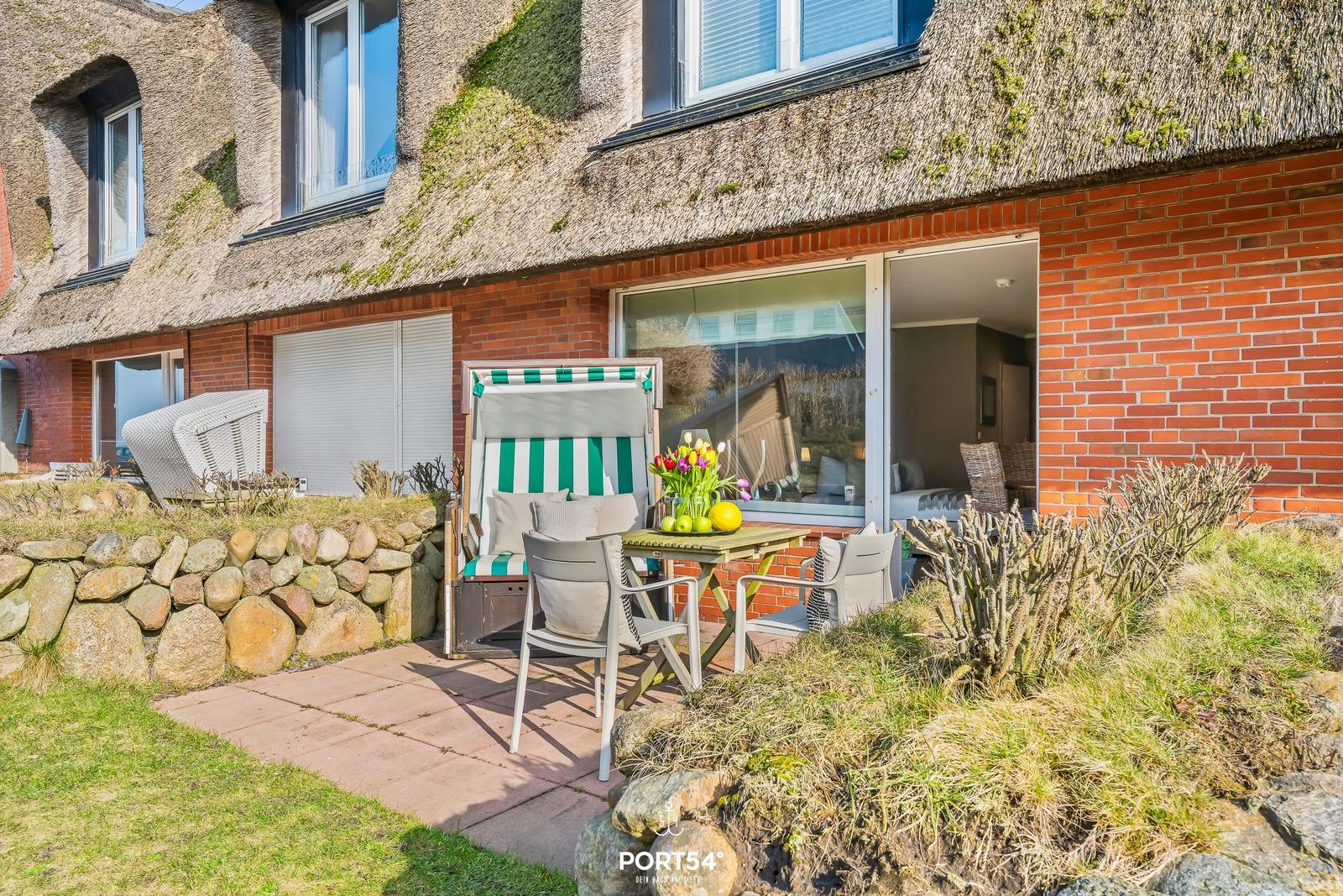 Terrace with lounge chair and table in front of a house with thatched roof and brick wall.