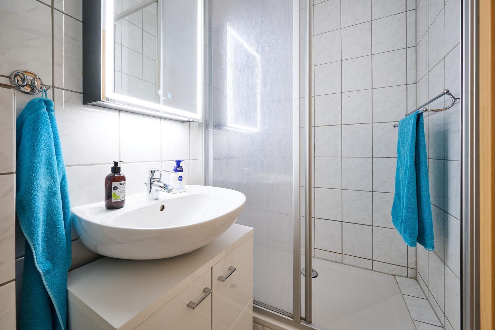 Badezimmer mit Waschbecken, Spiegel und Dusche. Blaue Handtücher hängen an Halterungen.