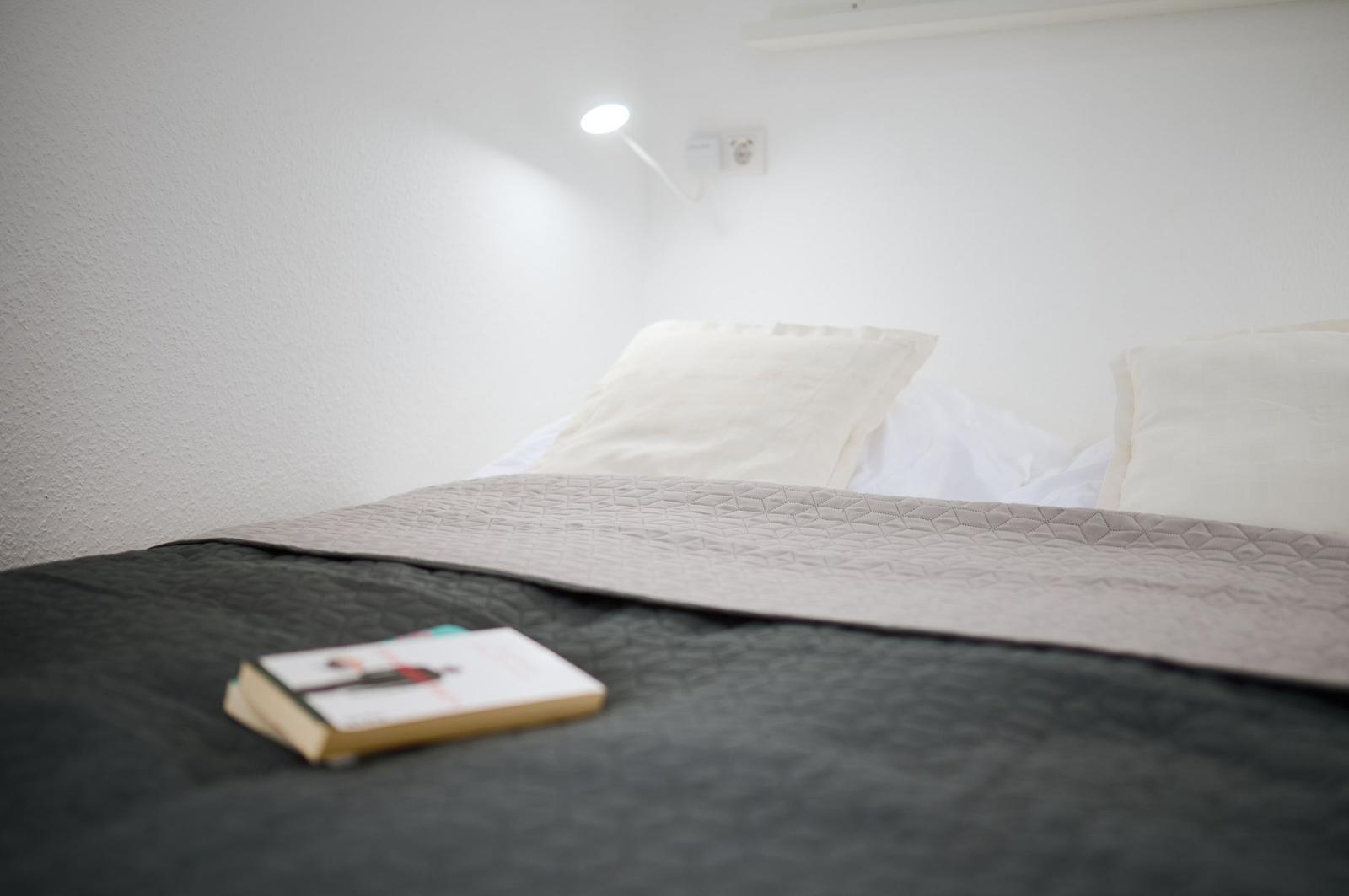 Ein Bett mit Kissen, einer Bettdecke und einem Buch. An der Wand hängt eine Lampe.