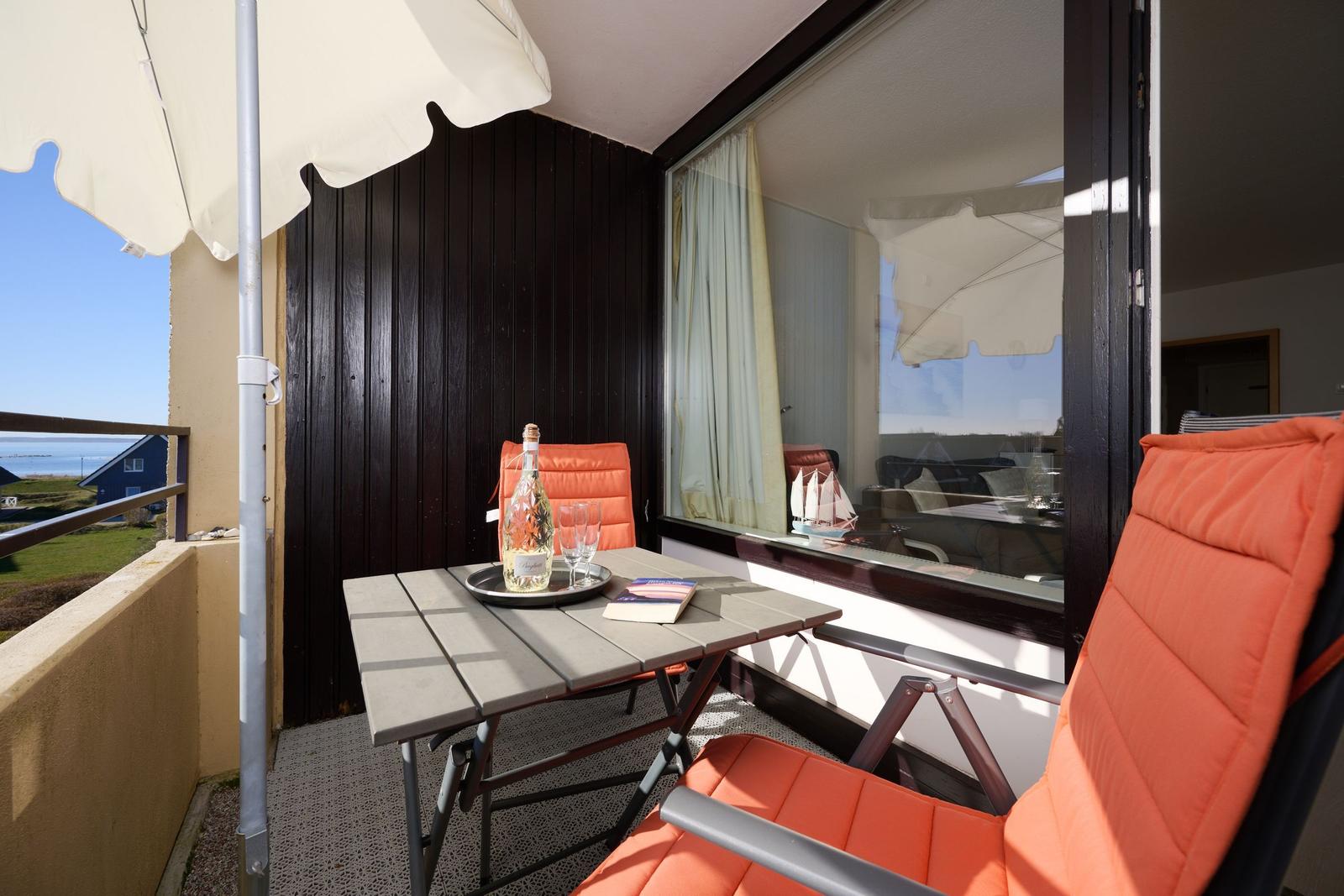Balkon mit Tisch, zwei orangefarbenen Stühlen und Sonnenschirm. Blick auf Küstenlandschaft.