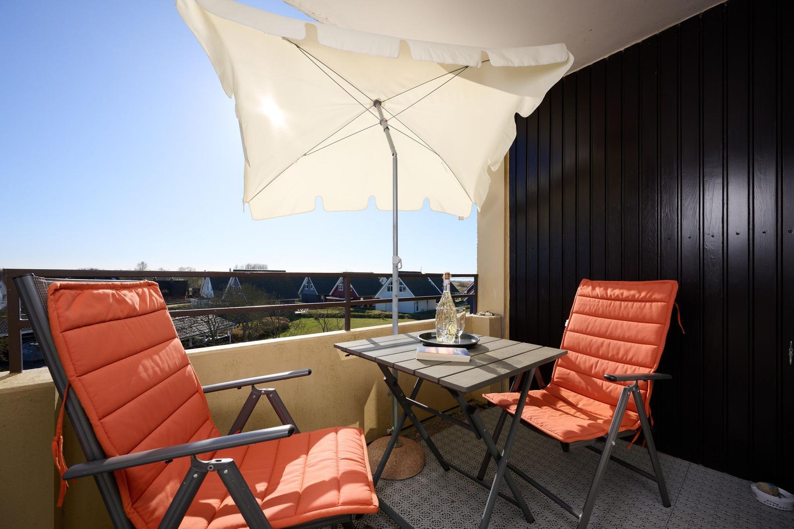 Terrasse mit zwei orangefarbenen Stühlen, Tisch und Sonnenschirm. Blick auf Dächer und Grünflächen.