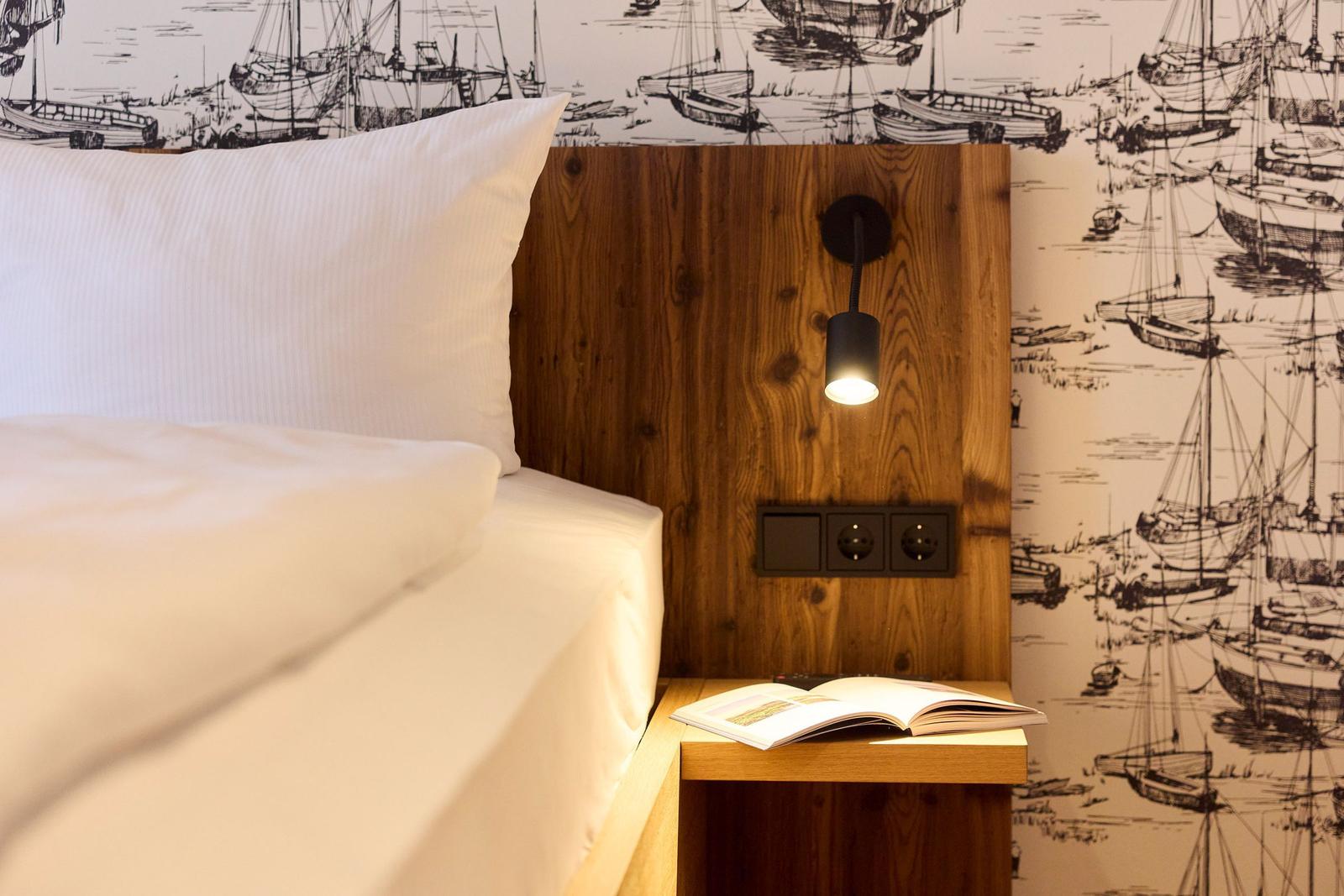 Schlafzimmer mit Holzkopfteil, Wandbeleuchtung und Segelyacht-Muster-Wandtapete.