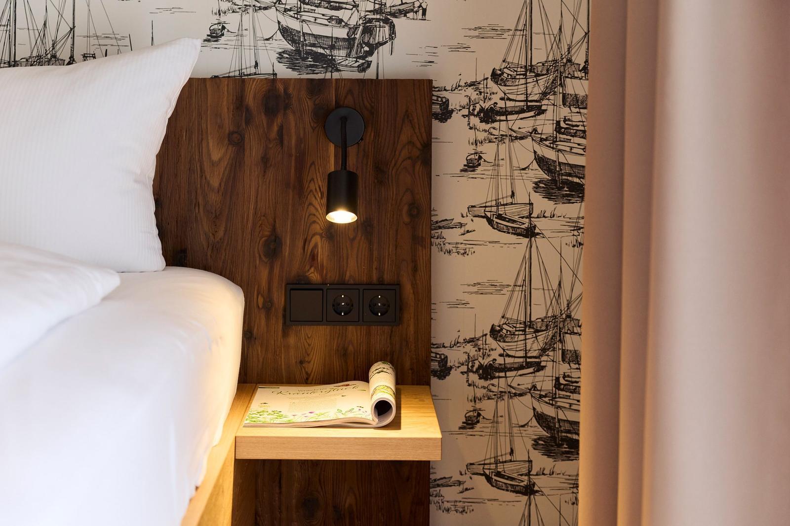 Bettkopf mit Holz, Wand mit Segelyachten-Motiv, Nachttisch mit Zeitschrift und Lampe.