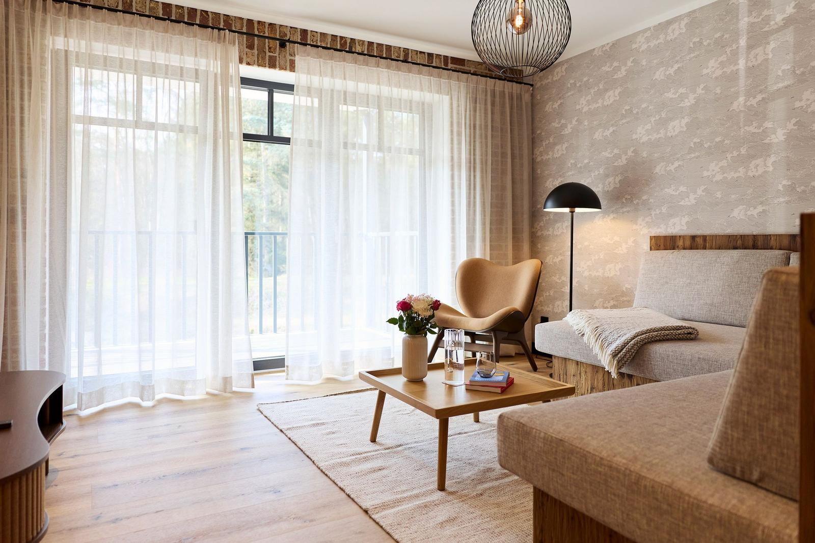 Wohnraum mit Sofa, Sessel, Tisch und Balkon. Holzboden und Fenster mit Vorhängen.