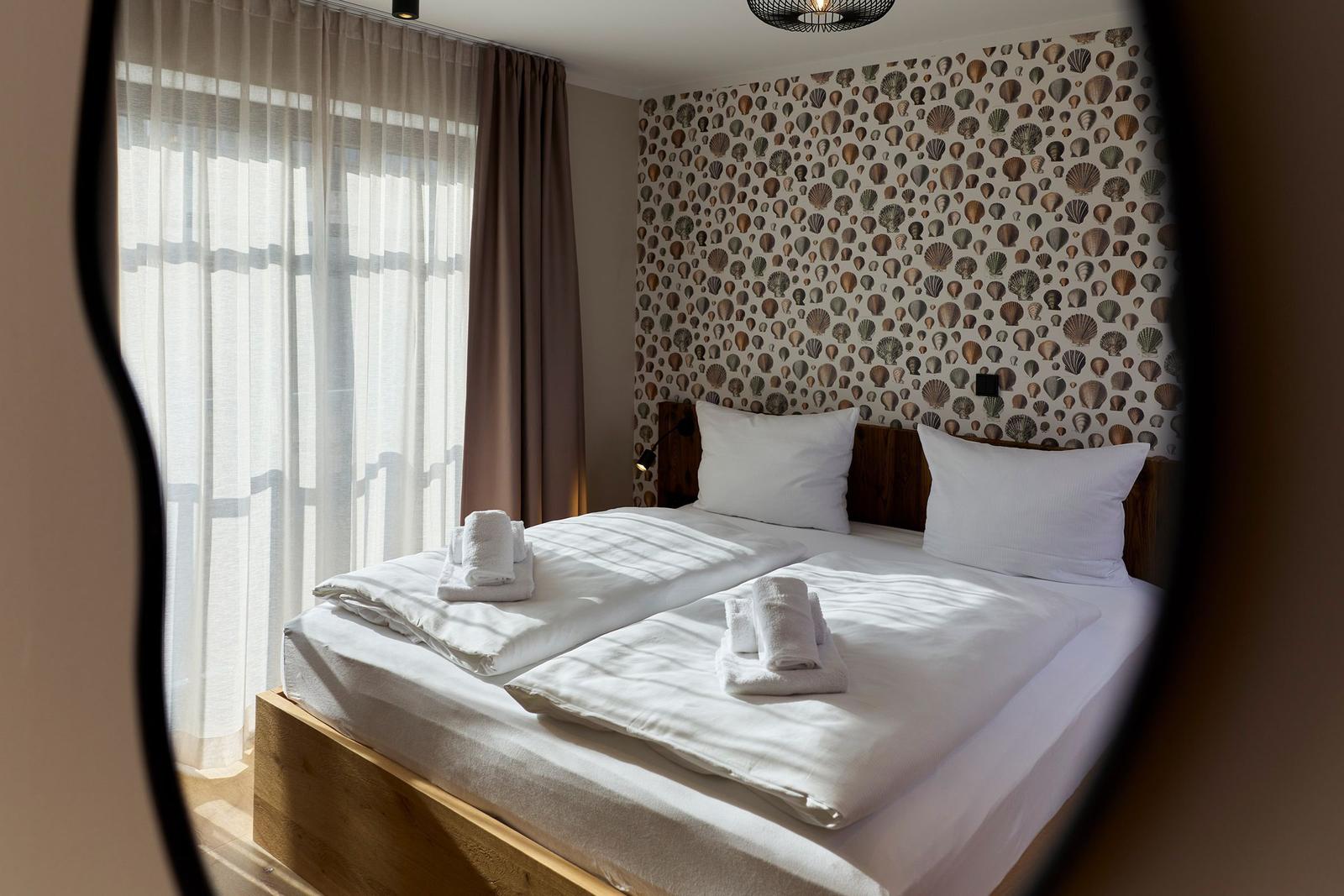 Schlafzimmer mit Bett, weißen Laken und Musterwand. Fenster mit Vorhängen.