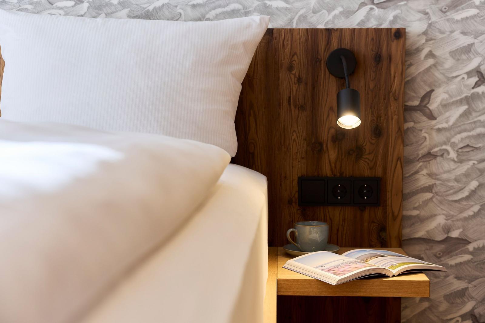 Schlafzimmer mit Holzkopfteil, Nachttisch, Lampe und offener Buch.