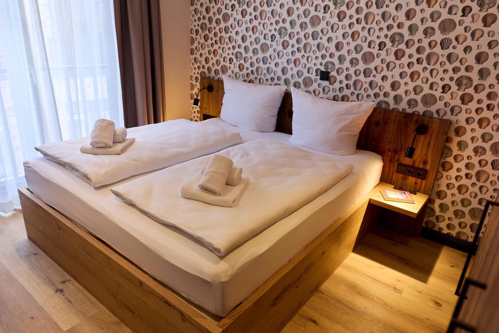 Doppelbett mit weißen Laken und Handtüchern. Hinter dem Kopfteil Wand mit Muschelmuster.