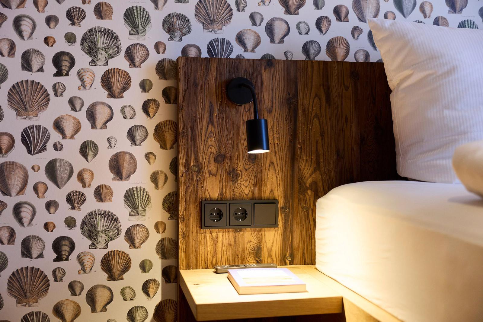 Schlafzimmer mit Holzkopfteil, Schalentapete und Nachttisch mit Buch.