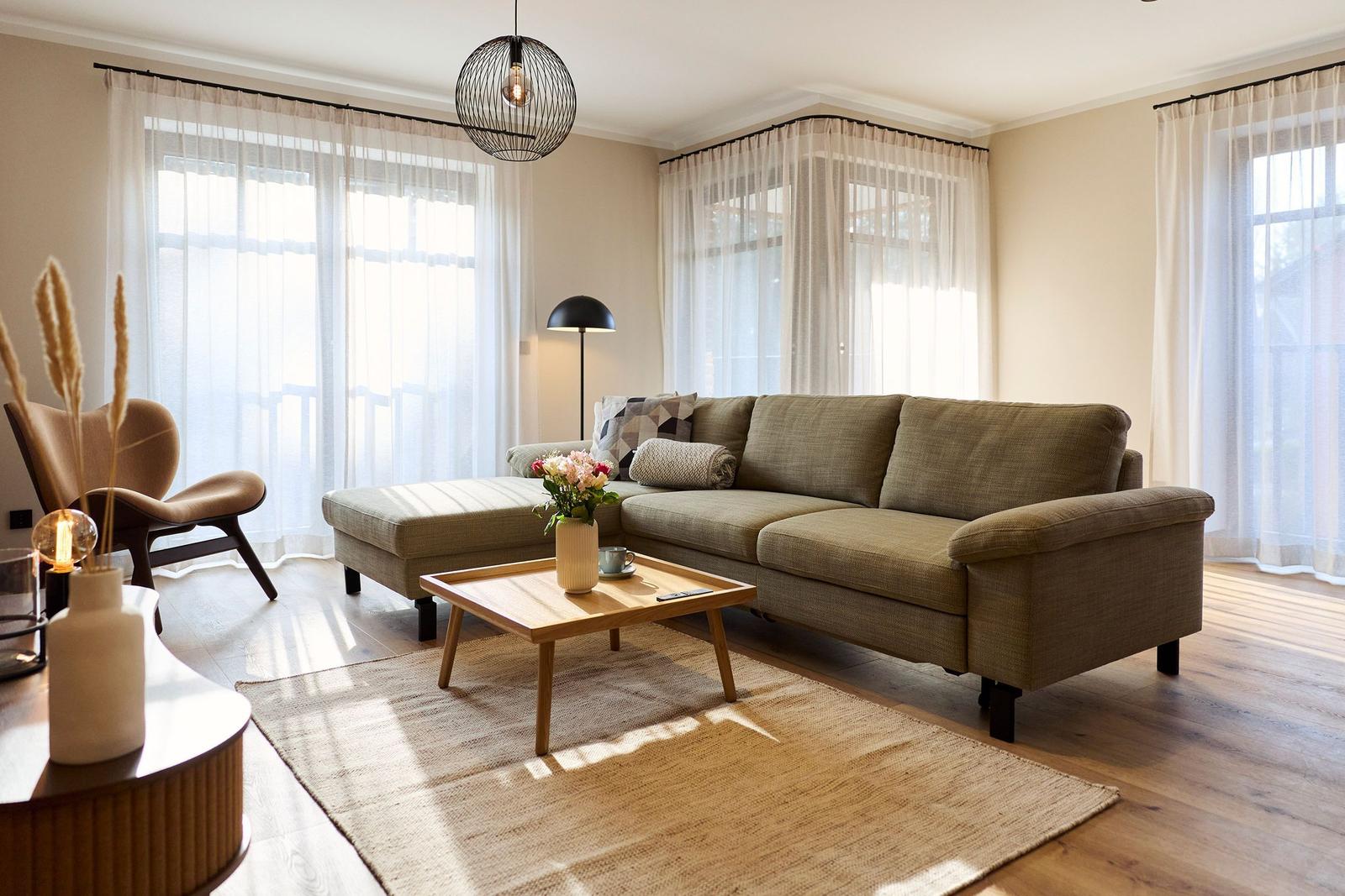 Wohnzimmer mit Sofa, Couchtisch und großen Fenstern mit Vorhängen.