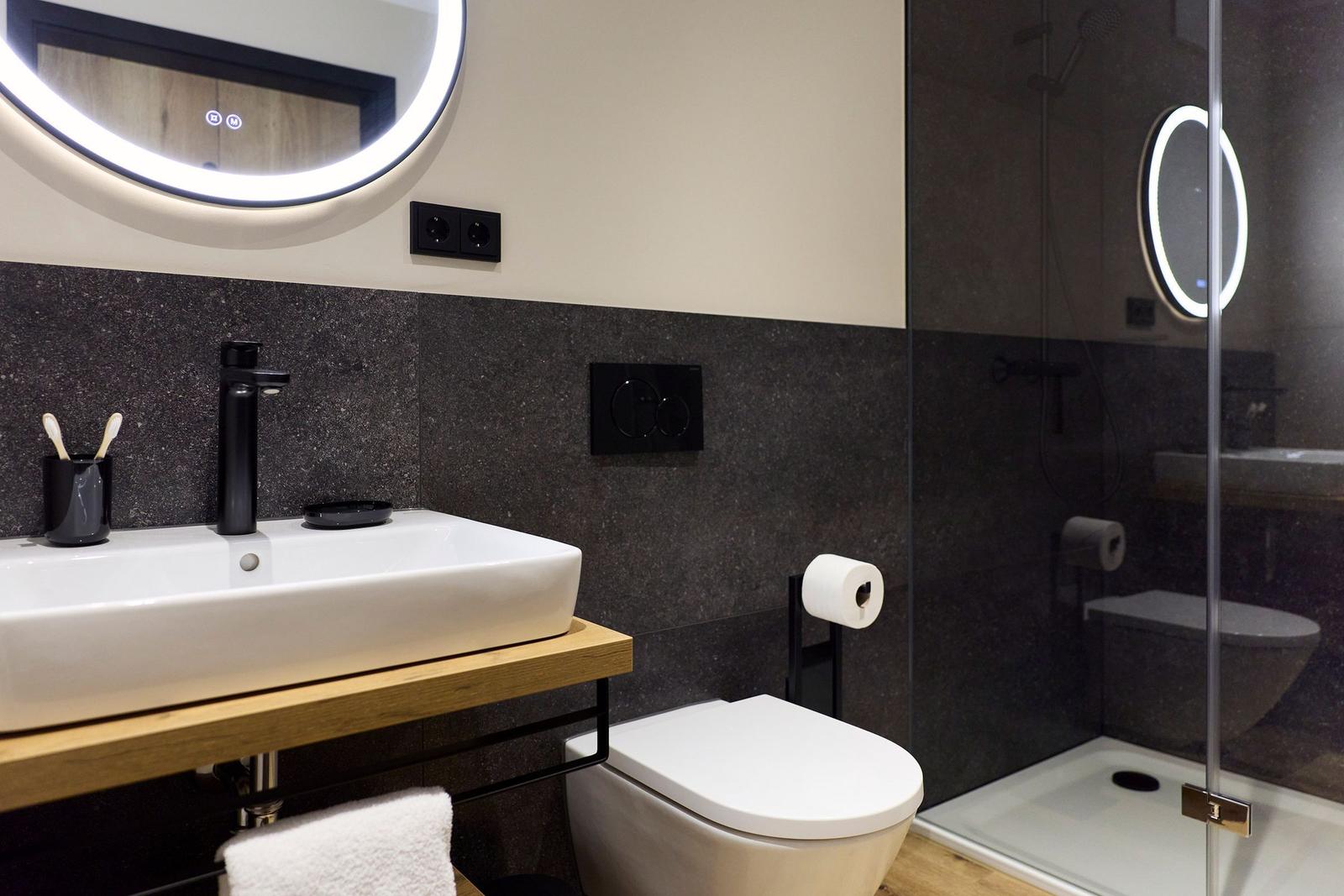 Badezimmer mit Waschbecken, Toilette und Dusche. Schwarze Wandfliesen und runder Spiegel.