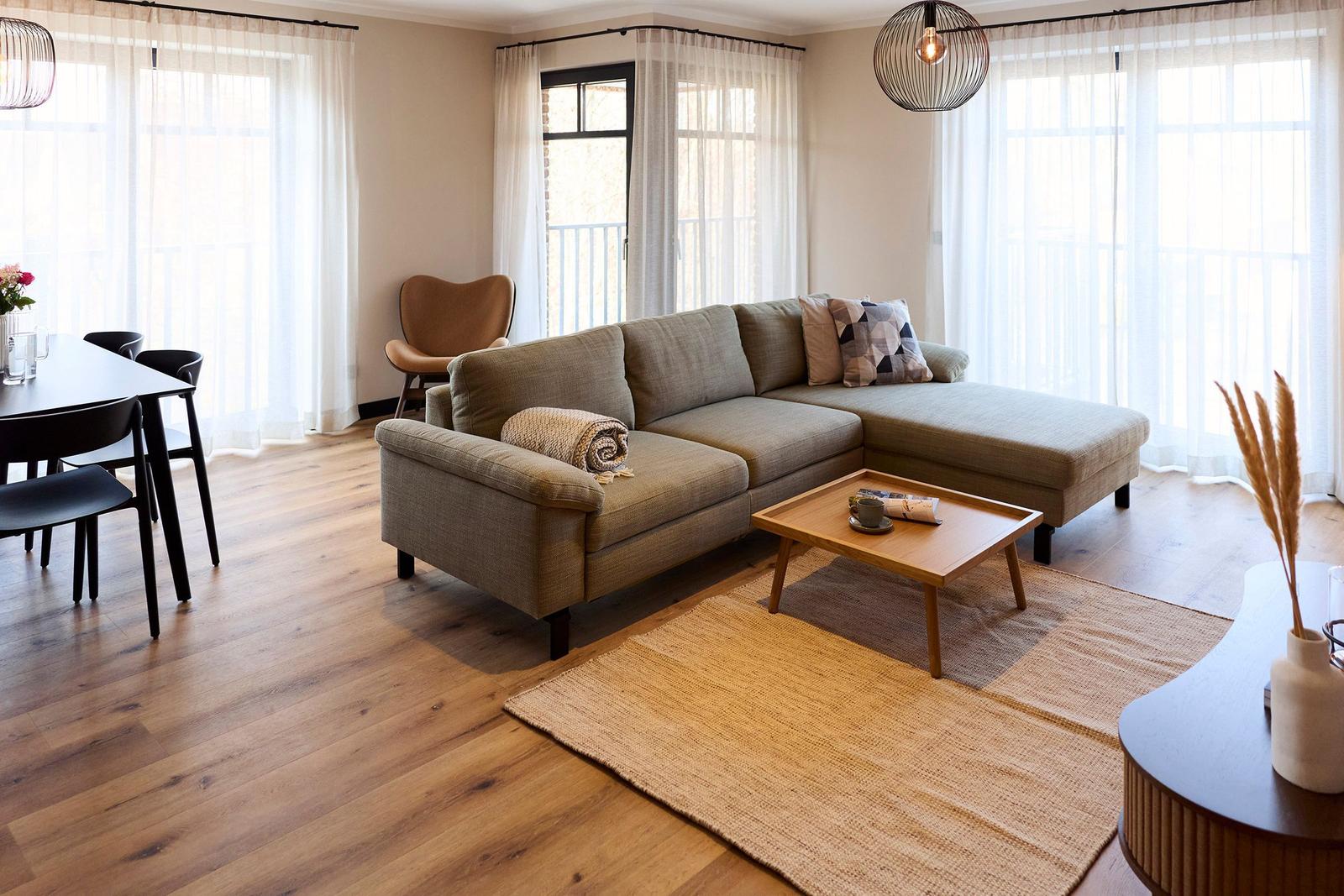 Wohnbereich mit Sofa, Tisch und Fenstern. Holzboden und helle Vorhänge.