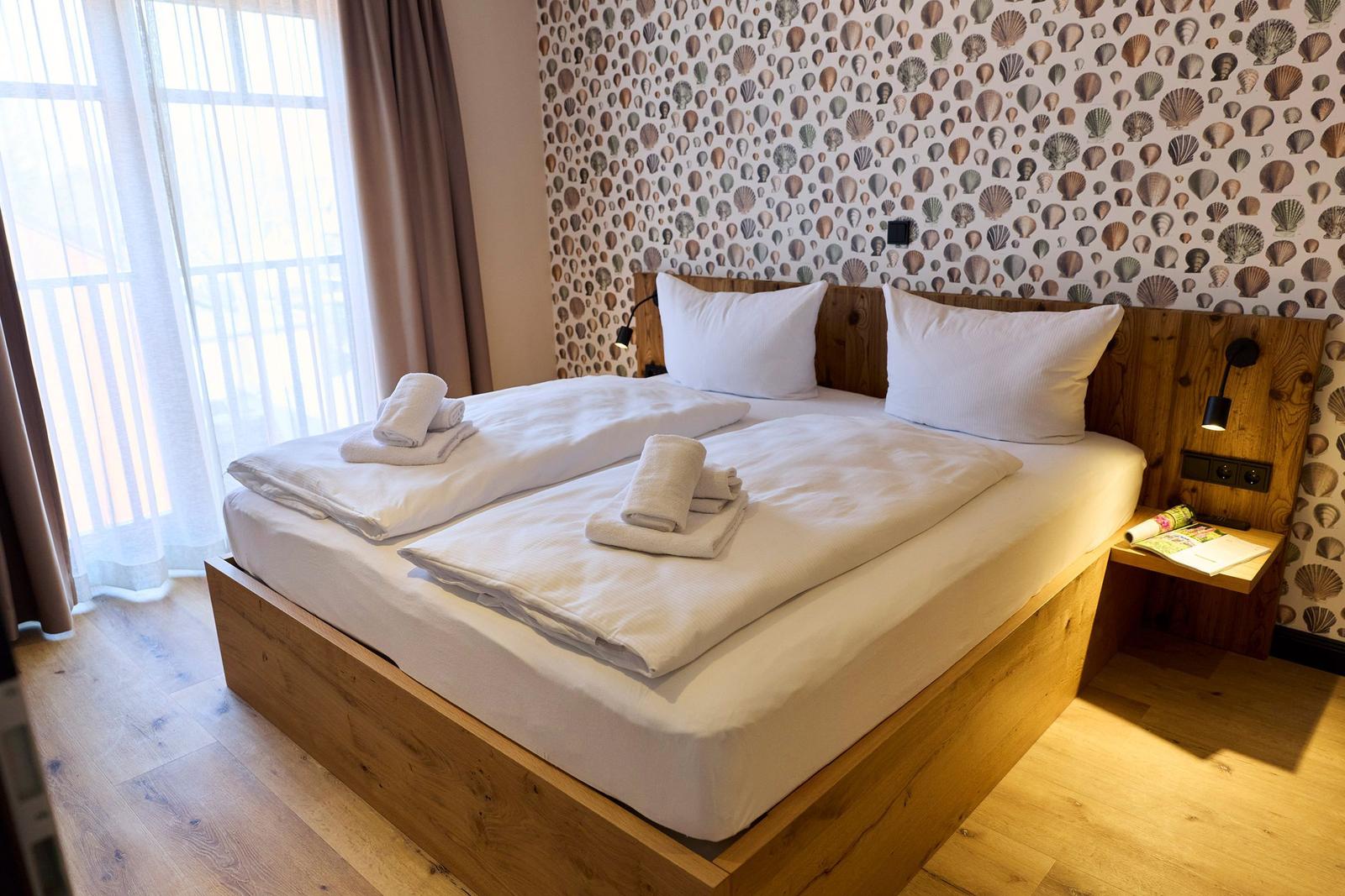 Doppelbett mit weißer Bettwäsche, Holzrahmen und Schalentapete an der Wand.