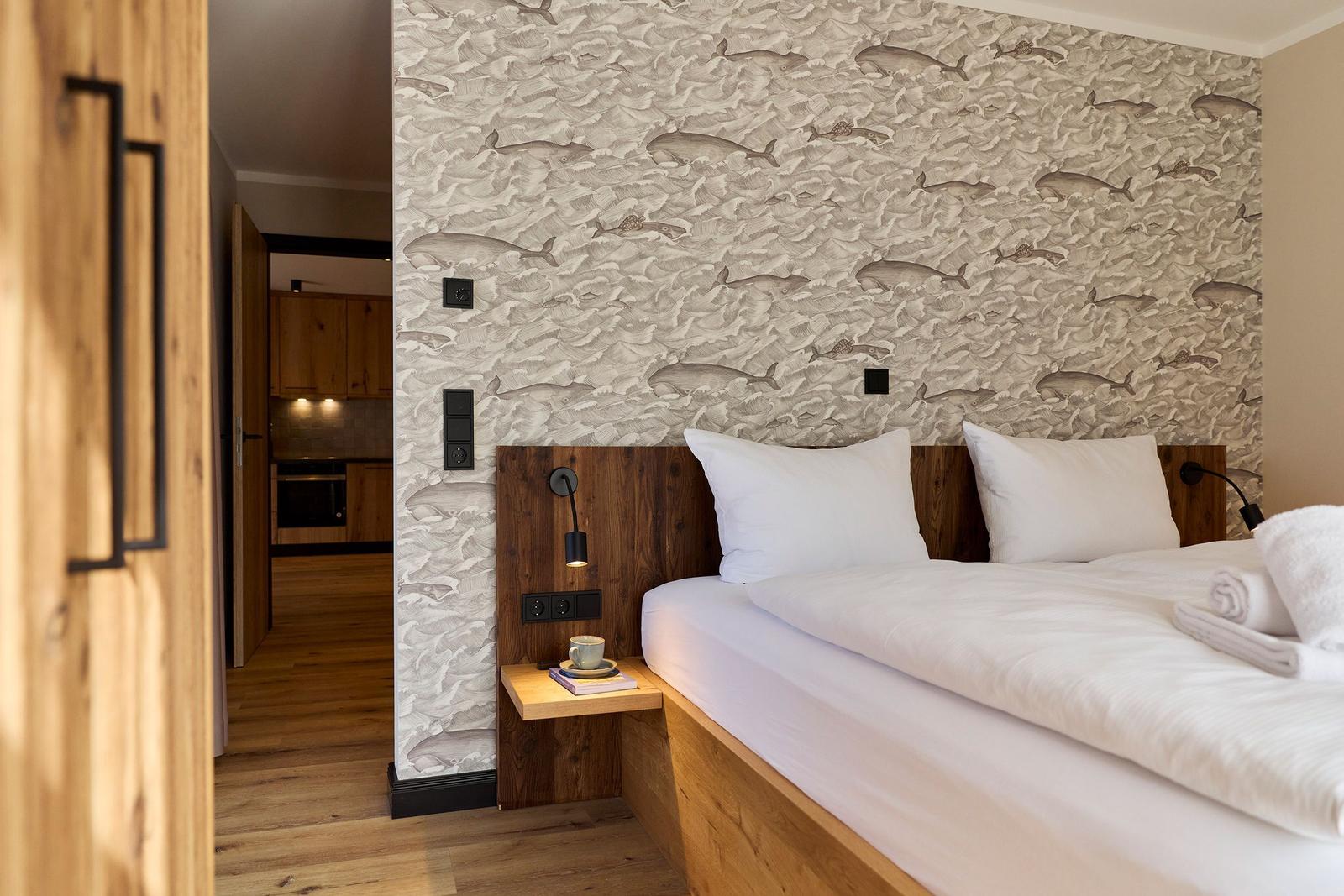 Schlafzimmer mit Bett, Holzgestell und Wand mit Wal-Motiv. Nebenraum mit Küchenmöbeln sichtbar.