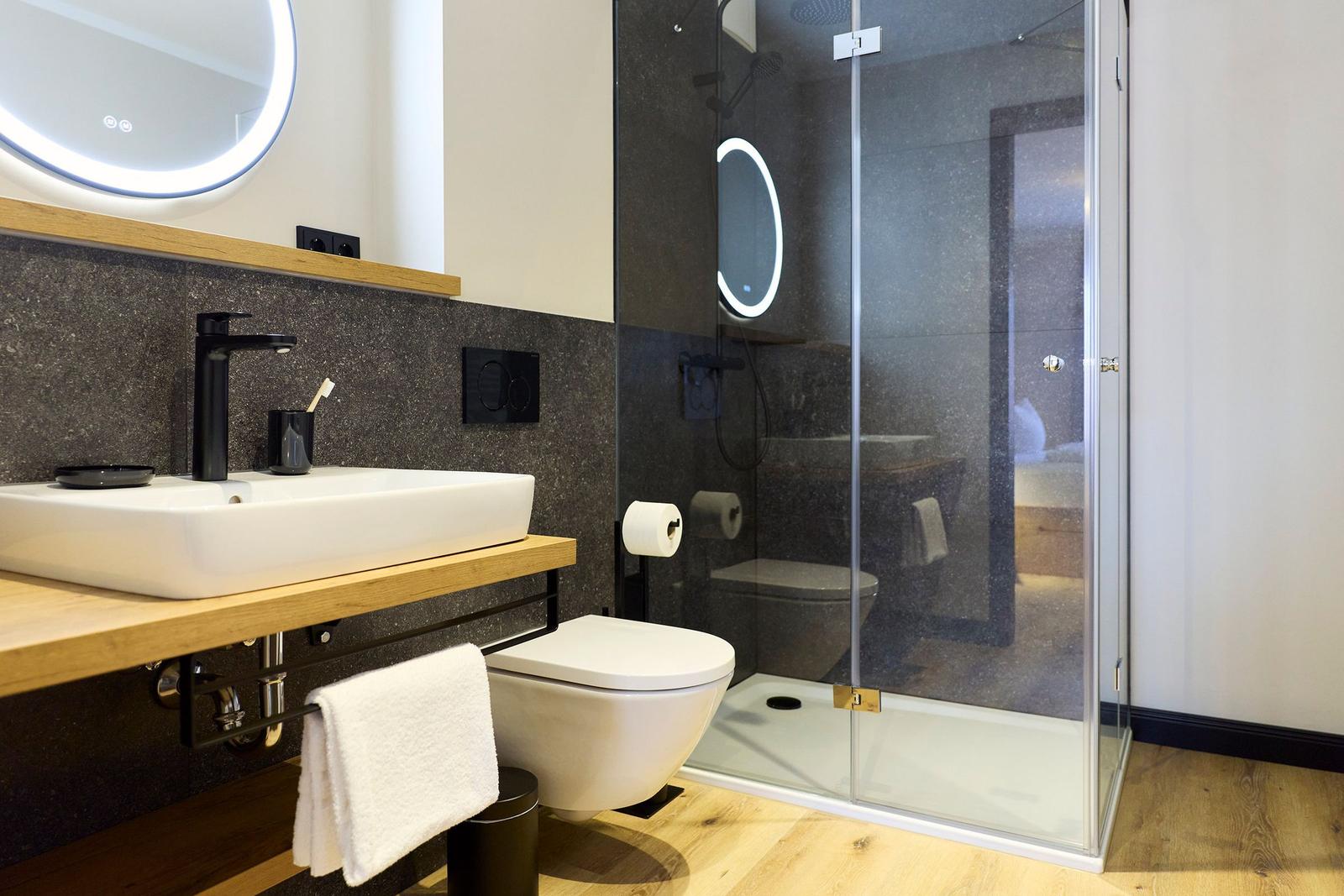 Badezimmer mit Dusche, Waschbecken und Toilette. Holzoberfläche und dunkle Wandfliesen.