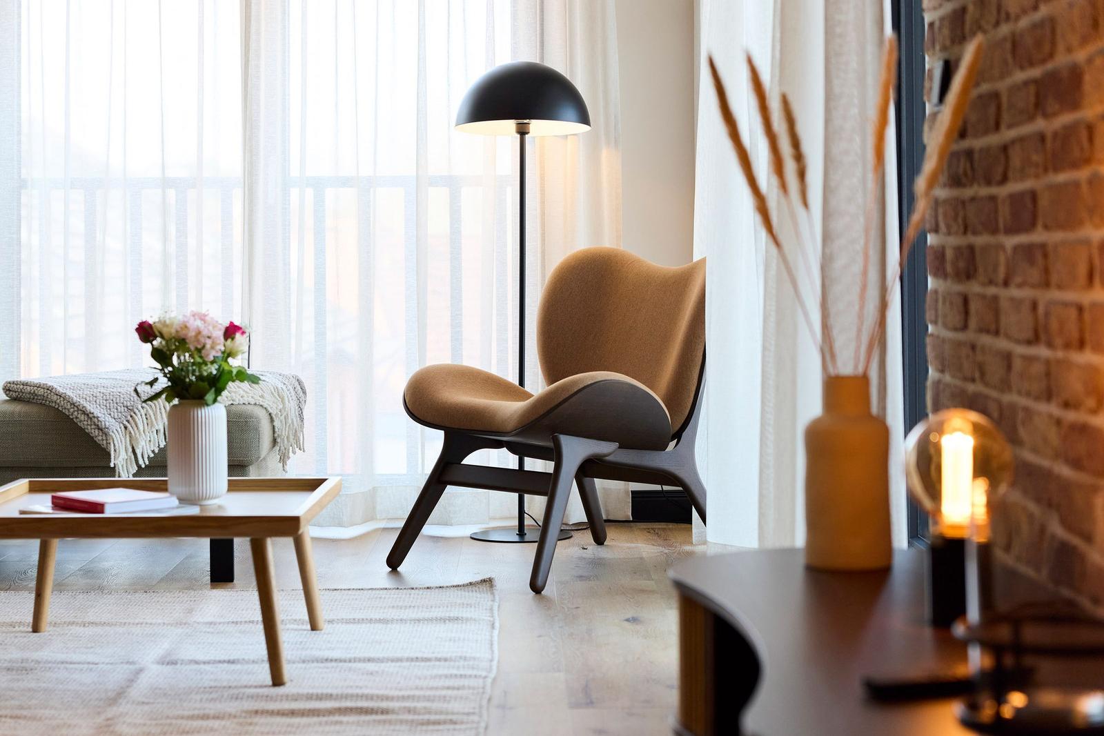 Ein Wohnbereich mit Sessel, Tisch, Lampe und Fenster mit Vorhängen.