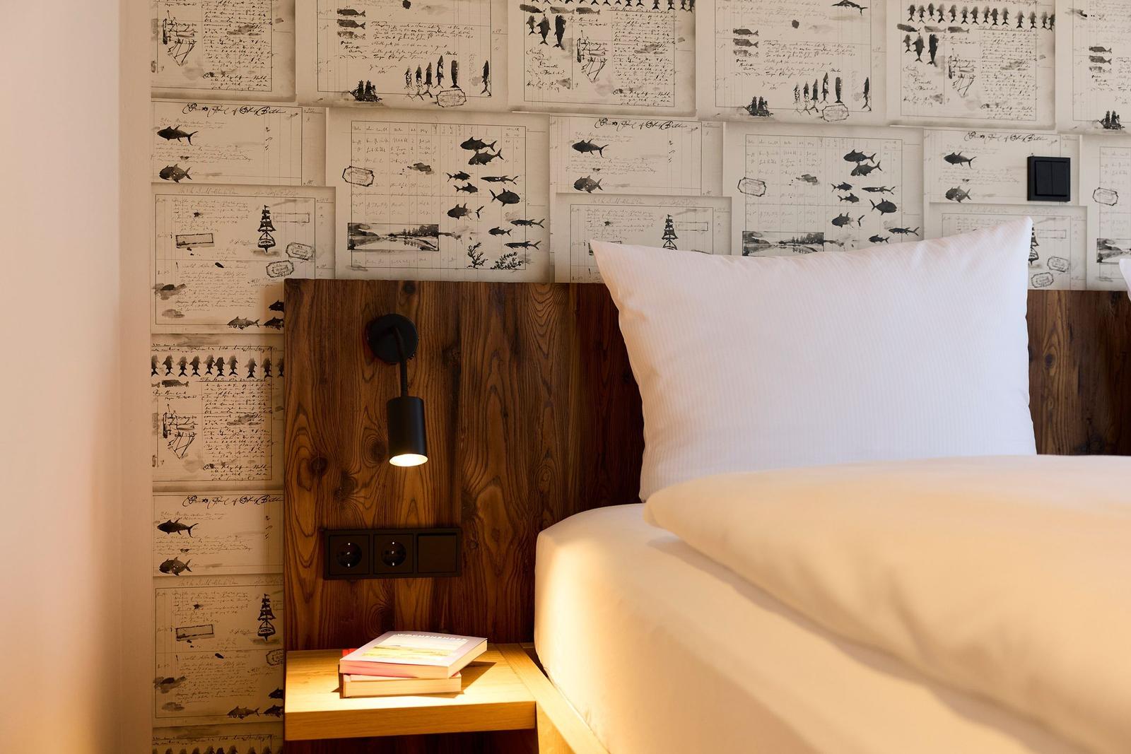 Schlafzimmer mit Holzkopfteil, Wand mit Briefmotiven und Nachttisch mit Buch.