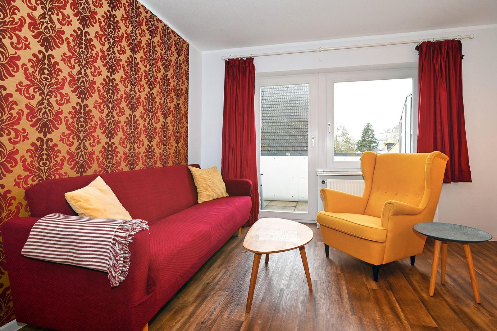 Wohnzimmer mit rotem Sofa, gelber Sessel und Holzboden. Wand mit Muster, Fenster mit roten Vorhängen.