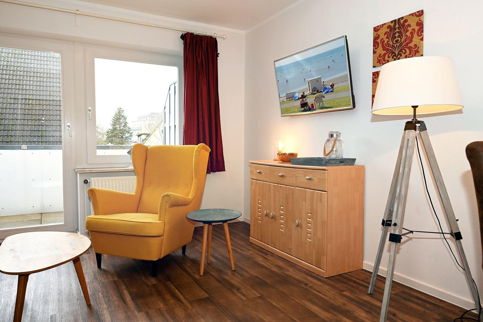 Ein Raum mit gelbem Sessel, Holzmöbeln und TV. Fenster mit rotem Vorhang.